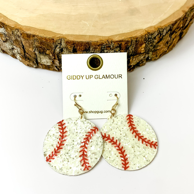 Pictured is circle glitter drop baseball earrings in white. They are propped up against a piece of circle wood on a white background.