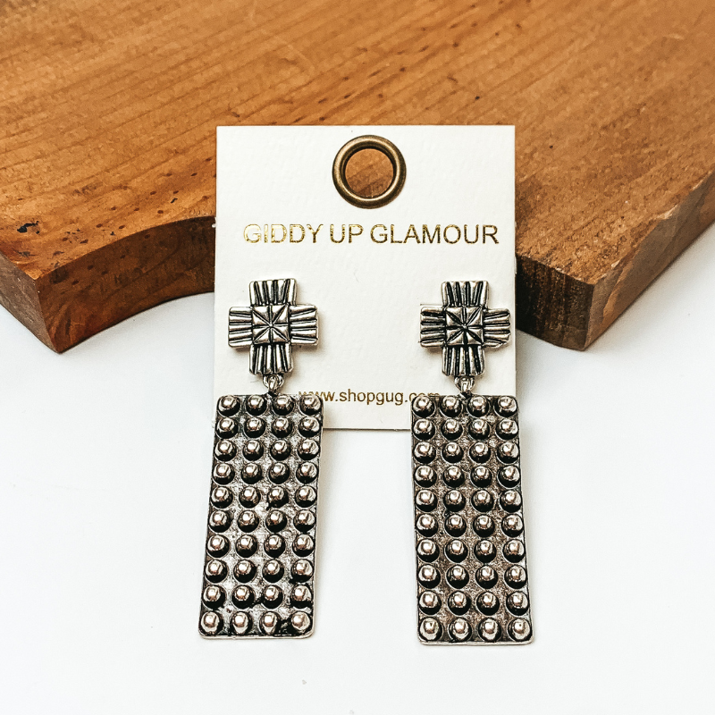 Pictured are rectangular concho drop earrings with a silver tone. They are propped up against a wooden board on a white background.