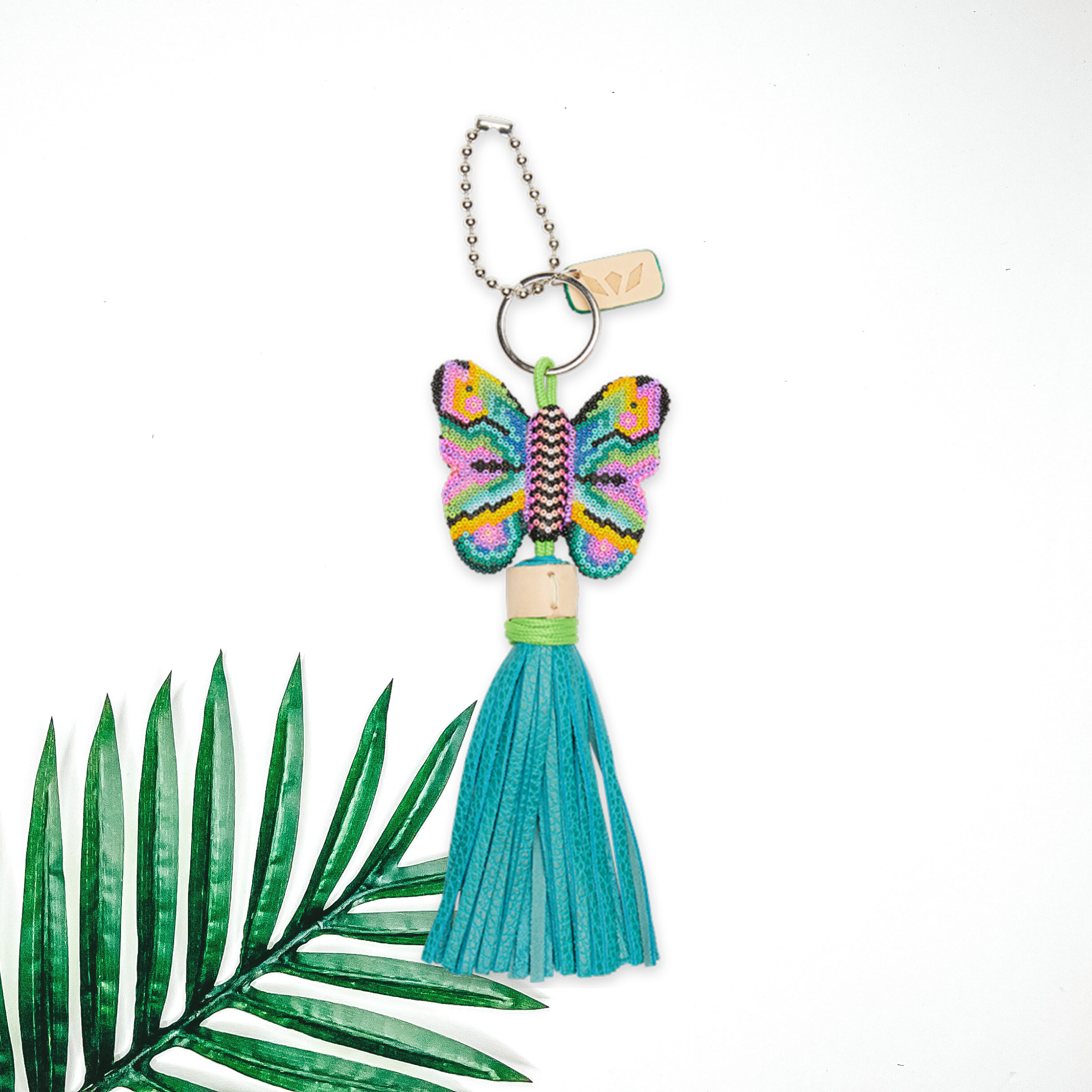 Centered in the middle of the picture is a butterfly charm with blue tassels. To the right of the charm is a palm leaf, all on a white background. 