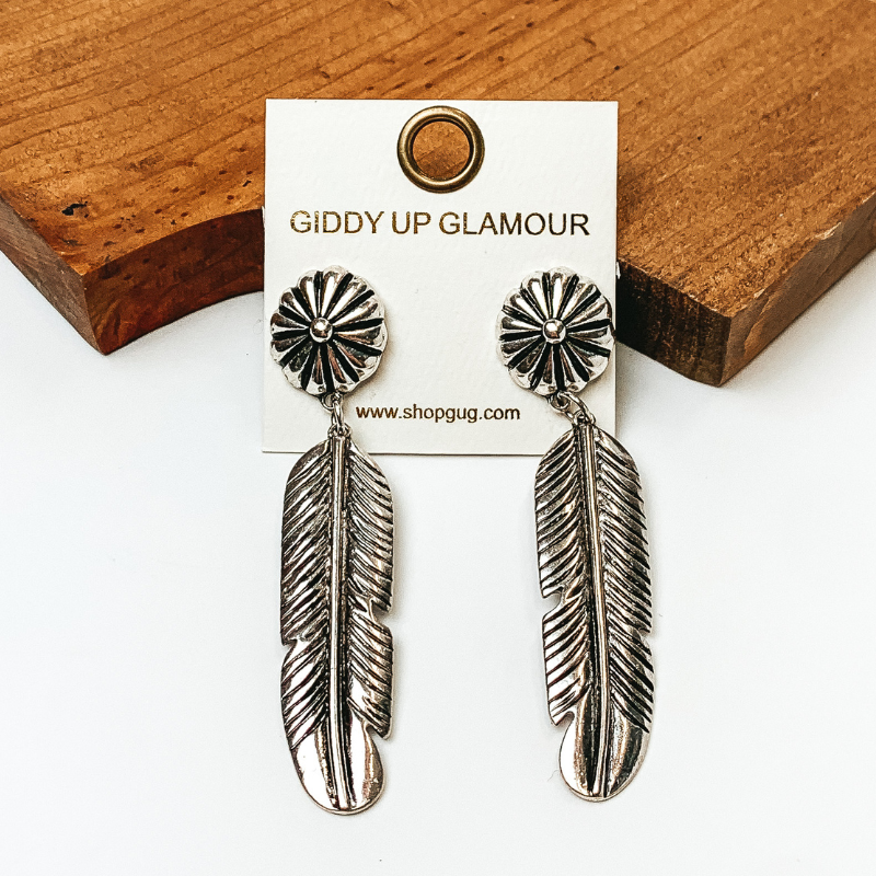 Pictured are feather drop earrings in silver with detailed studs. They are propped against a wooden board on a white background.