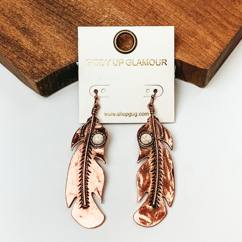 Pictured are copper feather drop earrings. They are propped on a wooden board on a white background.