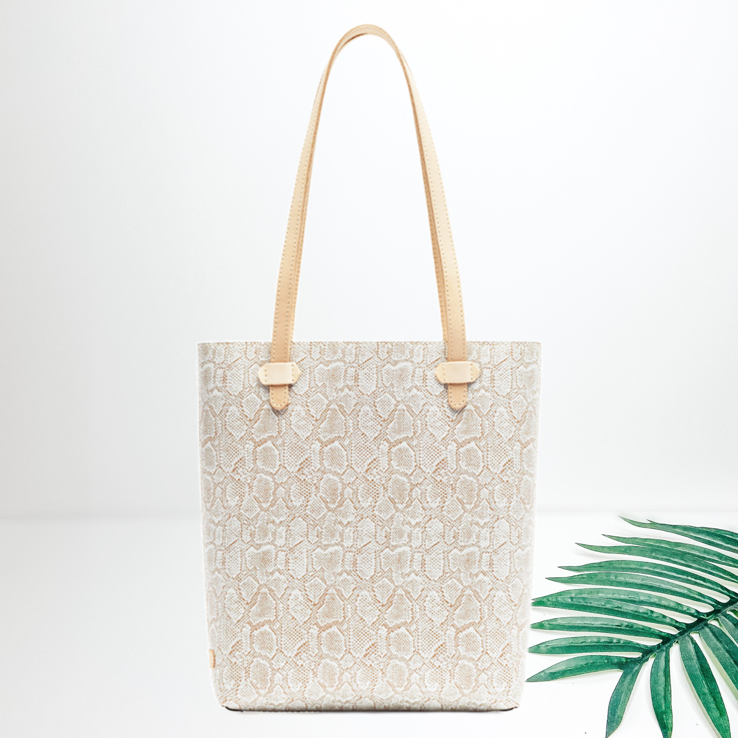Centered is a cream and white snakeskin tote. To the right of the tote is a palm leaf, all on a white background. 