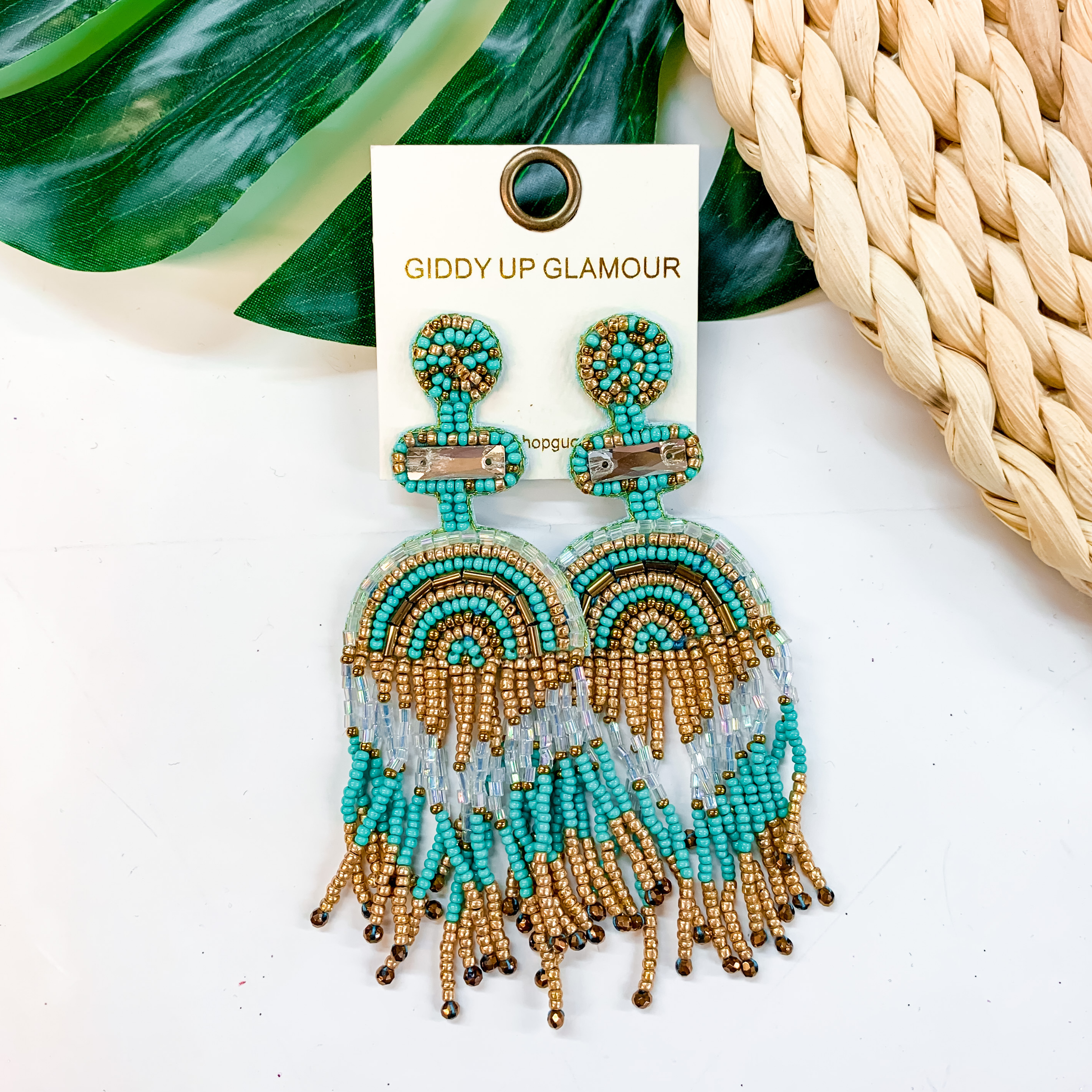 A pair of turquoise and gold beaded tassel earrings with a post back. These earrings are pictured on a white background with a basket weave and palm leaf.