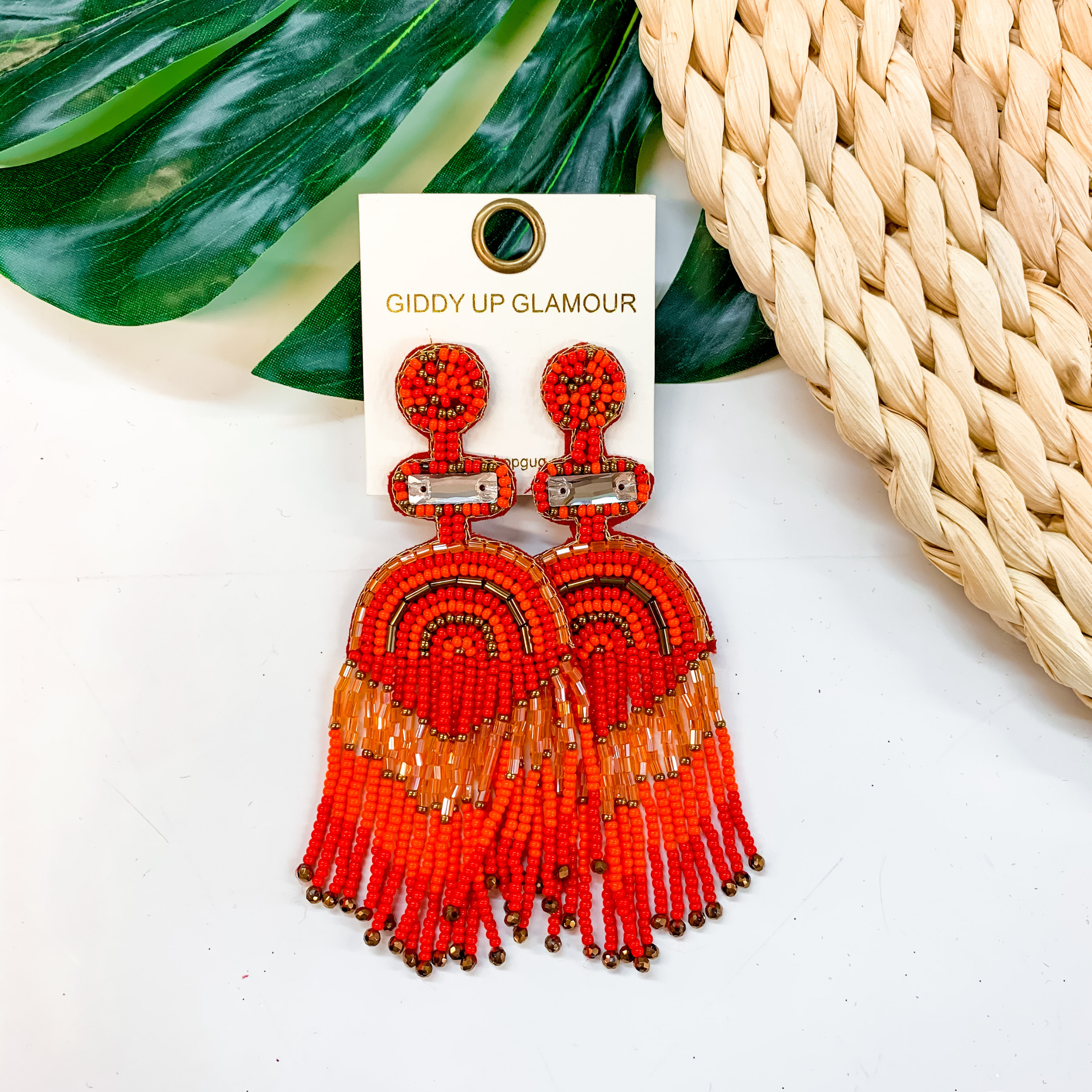 A pair of red beaded tassel earrings with a post back. These earrings are pictured on a white background with a basket weave and palm leaf.