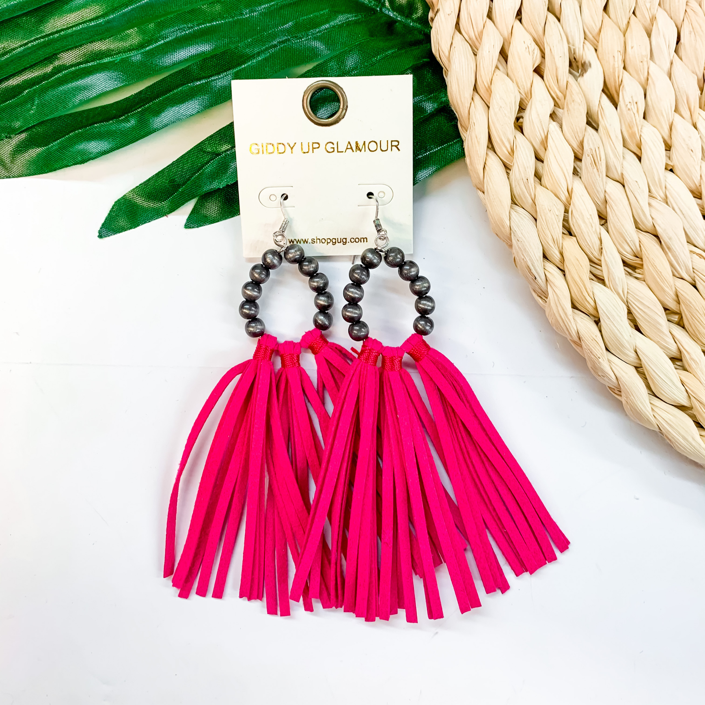 A pair of faux Navajo pearl earrings with hot pink tassels. These earrings are pictured on a white background with a basket and a green palm leaf.