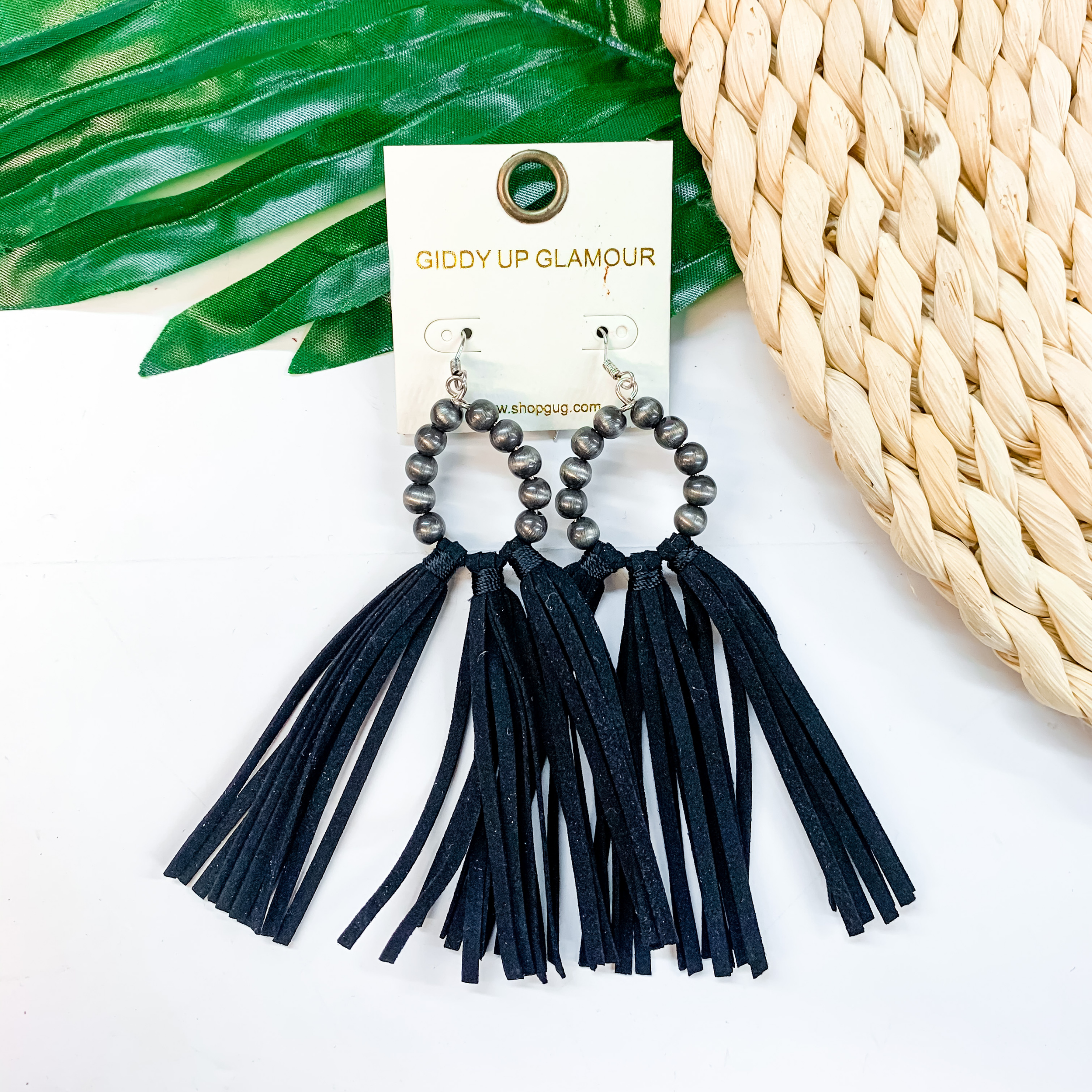 A pair of faux Navajo pearl earrings with black tassels. These earrings are pictured on a white background with a basket and a green palm leaf.