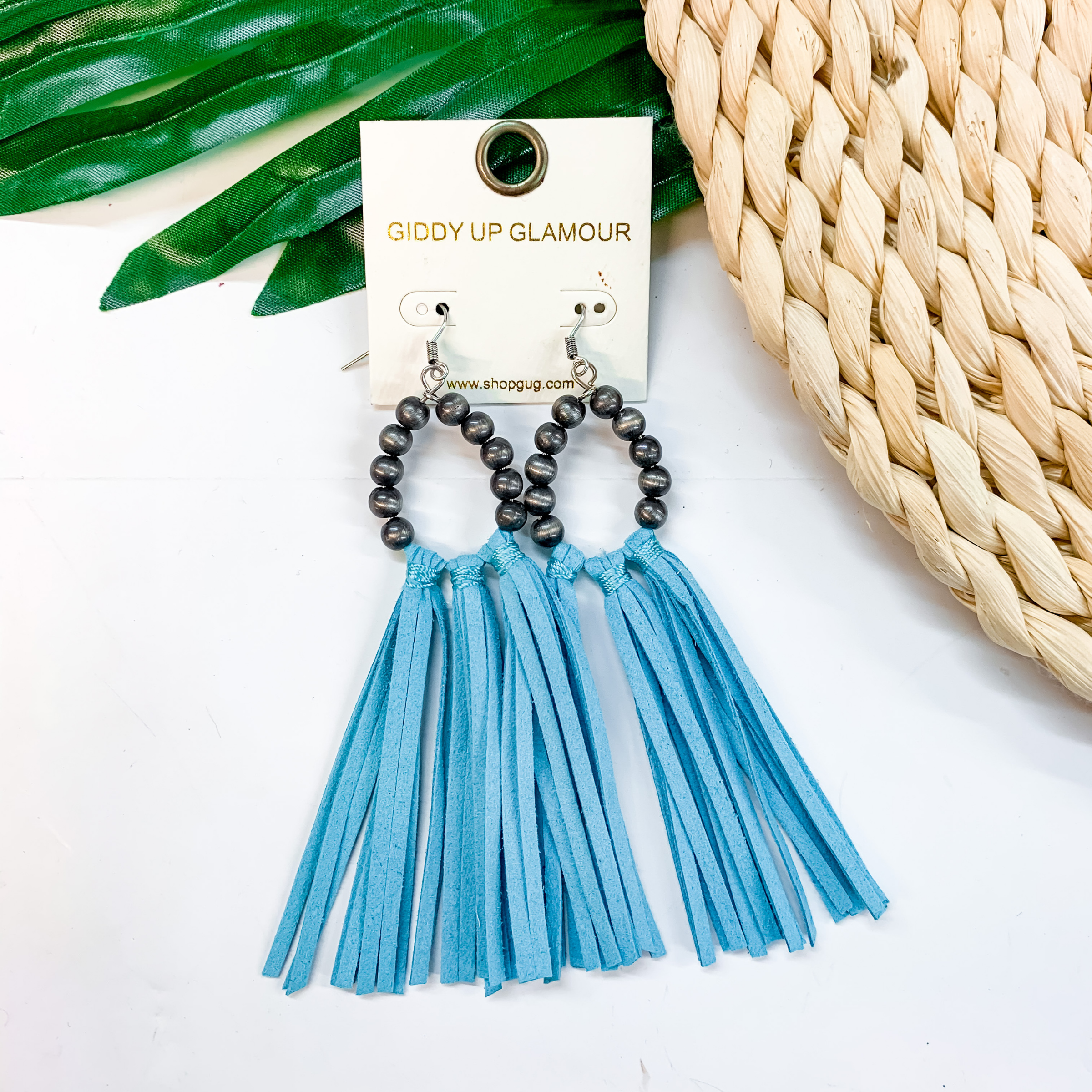 A pair of faux navajo pearl earrings with turquoise tassels. Pictured on a white background with a basket and palm leaf. 