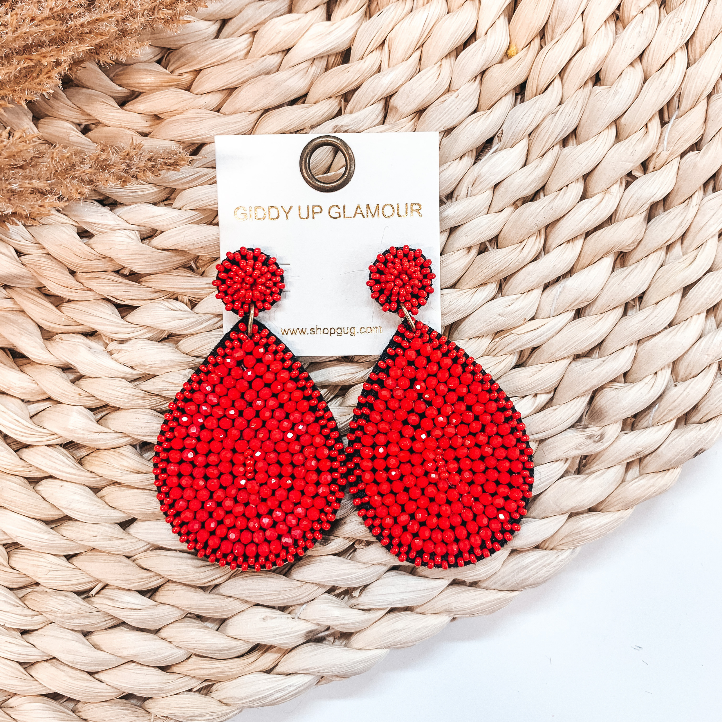 A pair of red crystal beaded teardrop earrings pictured on a basket weave display with pampus grass.