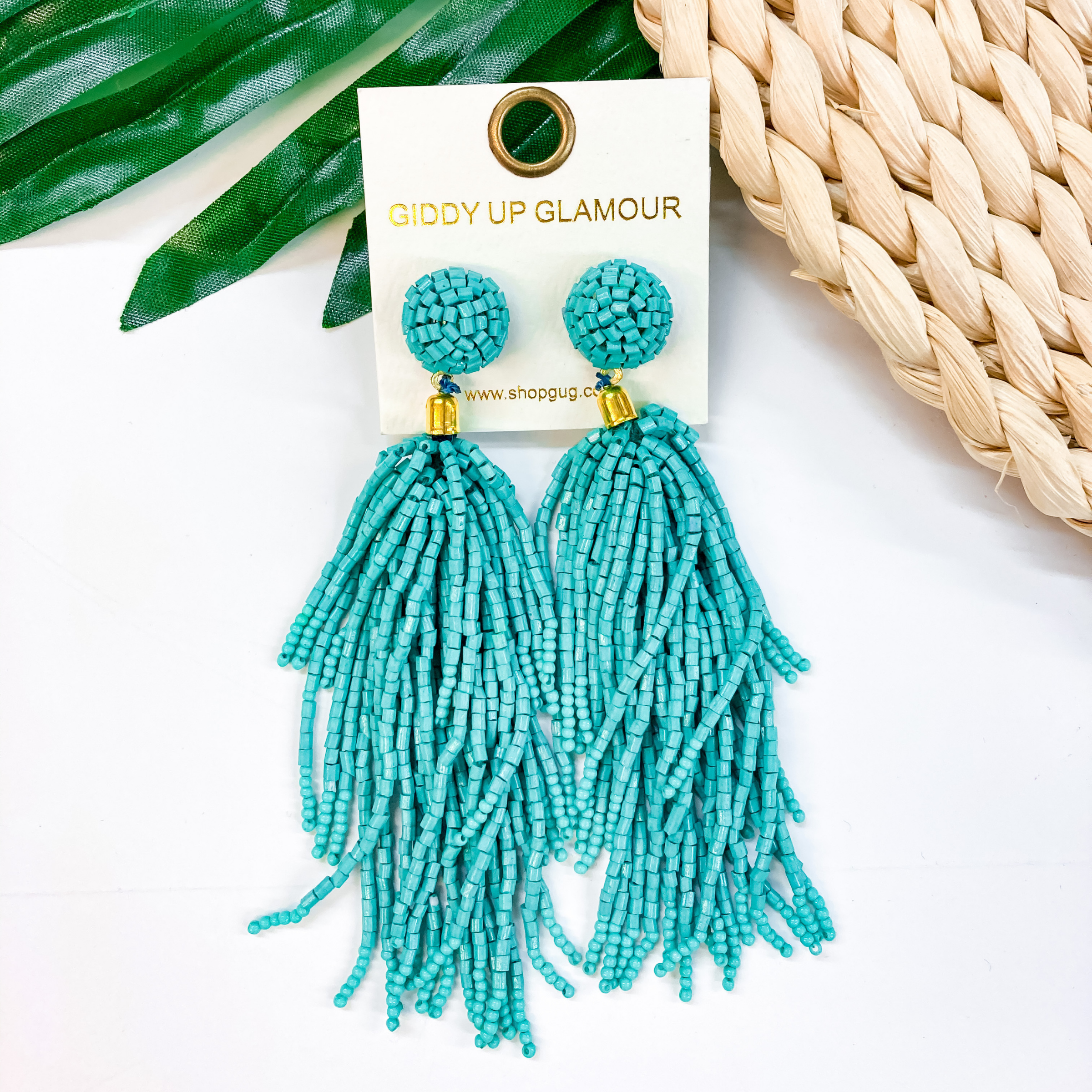 Circle, beaded post back stud earrings with a hanging beaded tassel in turquoise. These earrings are pictured on a white background with a green leaf and tan basket weave in the background. 