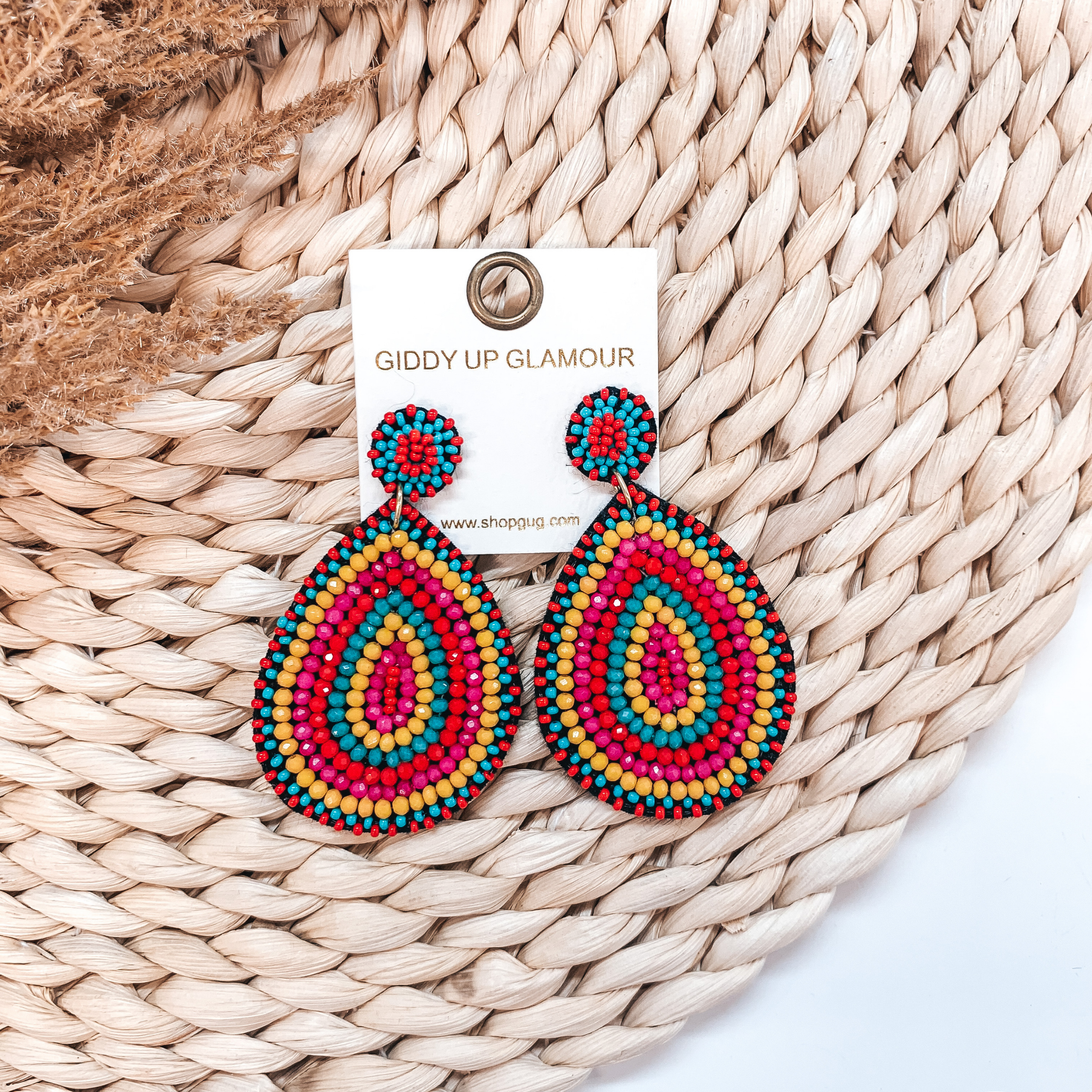 Circle, beaded post back stud earrings with a hanging, teardrop beaded pendant in multicolored. These earrings are pictured on a tan basket weave with a brown floral in the background.   