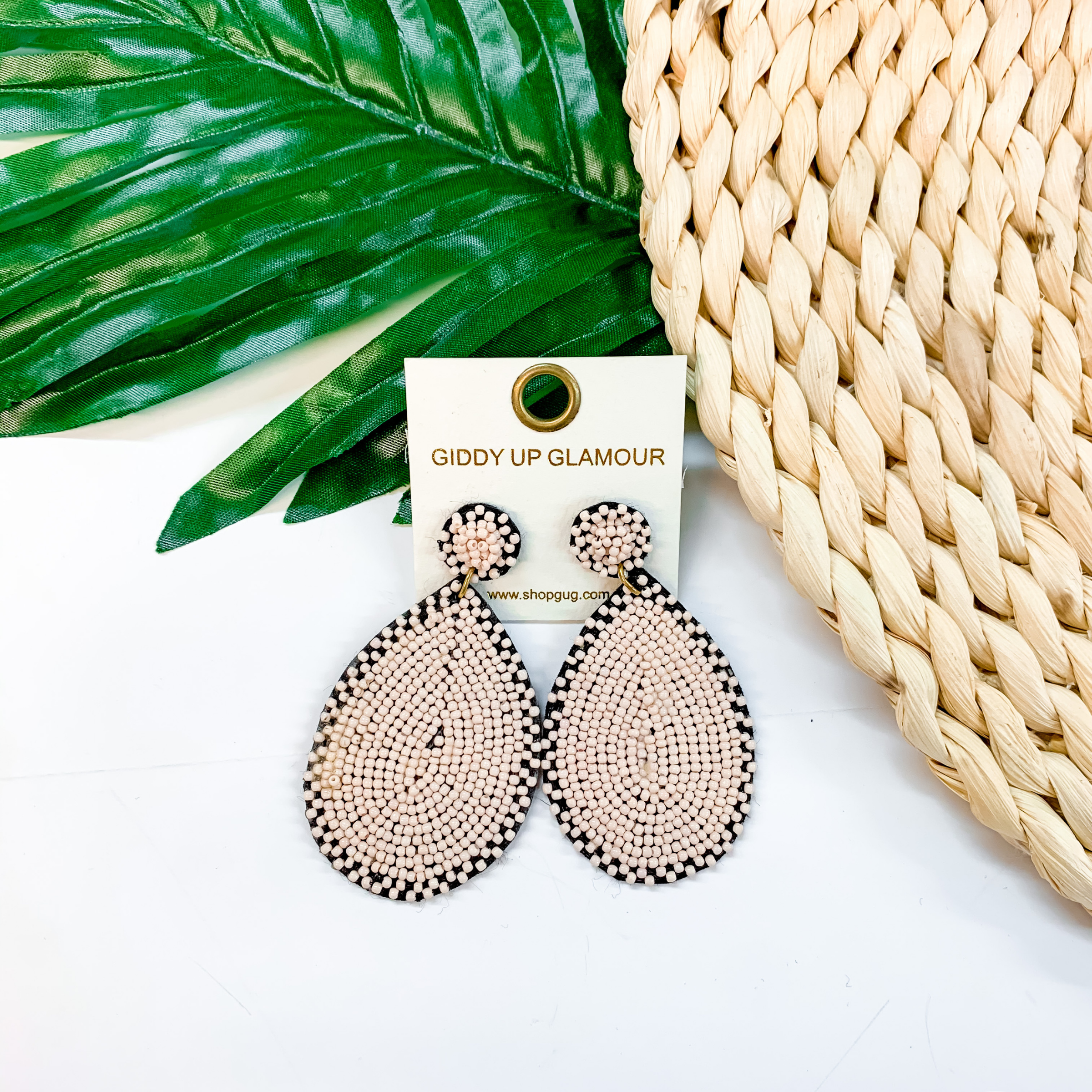 Circle, beaded post back stud earrings with a hanging, teardrop beaded pendant in blush pink. These earrings are pictured on a white background with tan basket weave with a green leaf in the background. 