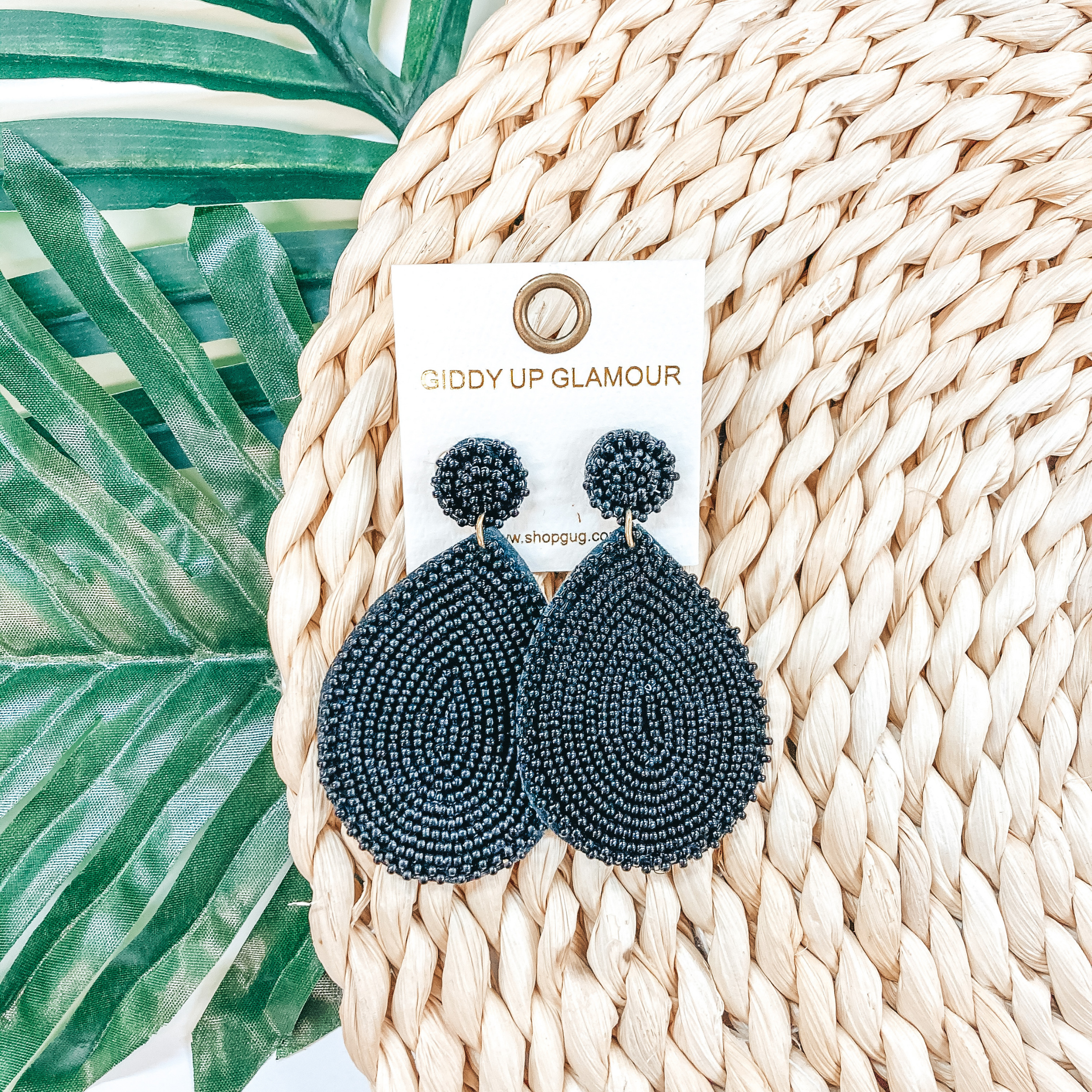 Circle, beaded post back stud earrings with a hanging, teardrop beaded pendant in black. These earrings are pictured on a tan basket weave with a green leaf in the background.