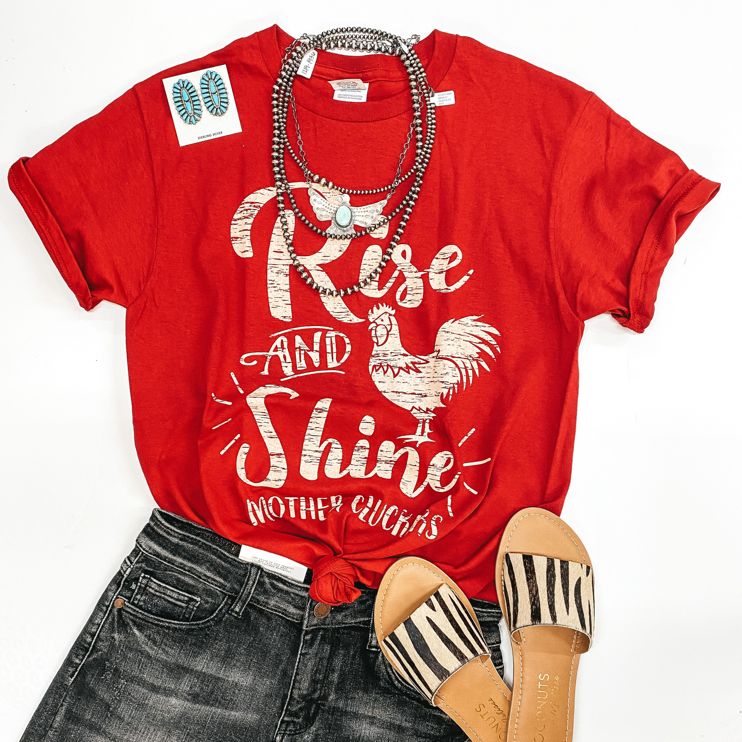 A red graphic tee that says " Rise and shine mother cluckers" with a rooster. Pictured with sandals, black shorts, and genuine Navajo earrings.