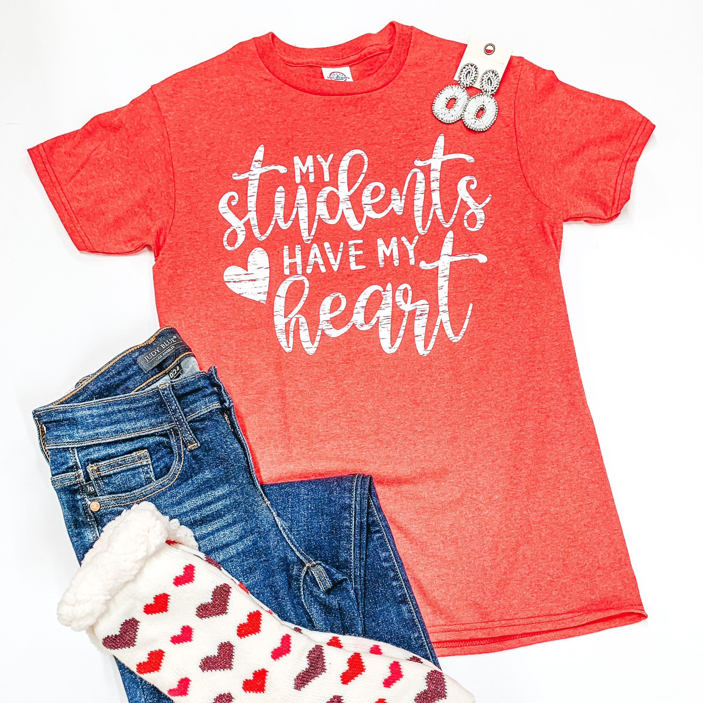 A red tee that says "my students have my heart"  is laid in the middle of the picture. Jeans are laid on the bottom left of the tee and heart socks are laid on top of the jeans. All is on a white background. 