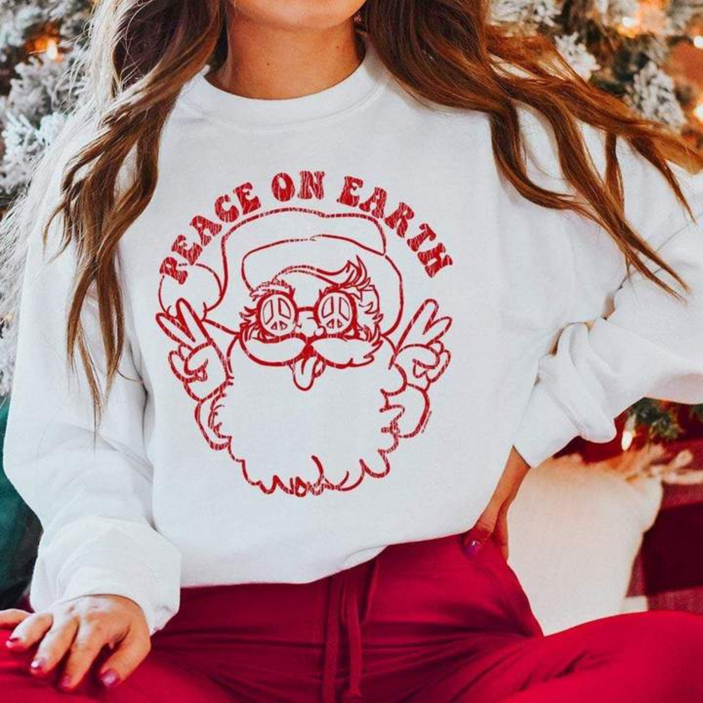 This white pullover includes a crew neckline, long sleeves, and a graphic that says "Peace on Earth" with a hippie santa all in red. The model has this sweatshirt modeled with a pair of red pants. 