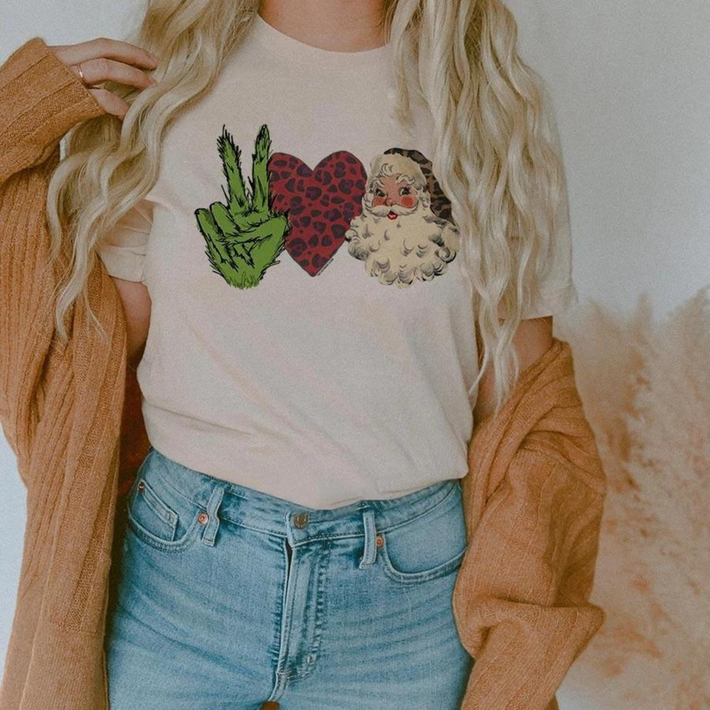 This cream tee includes a crew neckline, short sleeves, and a cute Christmas graphic of a Grinch hand throwing up a peace sign, a red leopard print heart, and a Santa Clause with a leopard hat side by side. The model has this graphic tee styled with rolled sleeves, a camel cardigan, and a light wash pair of denim jeans. 