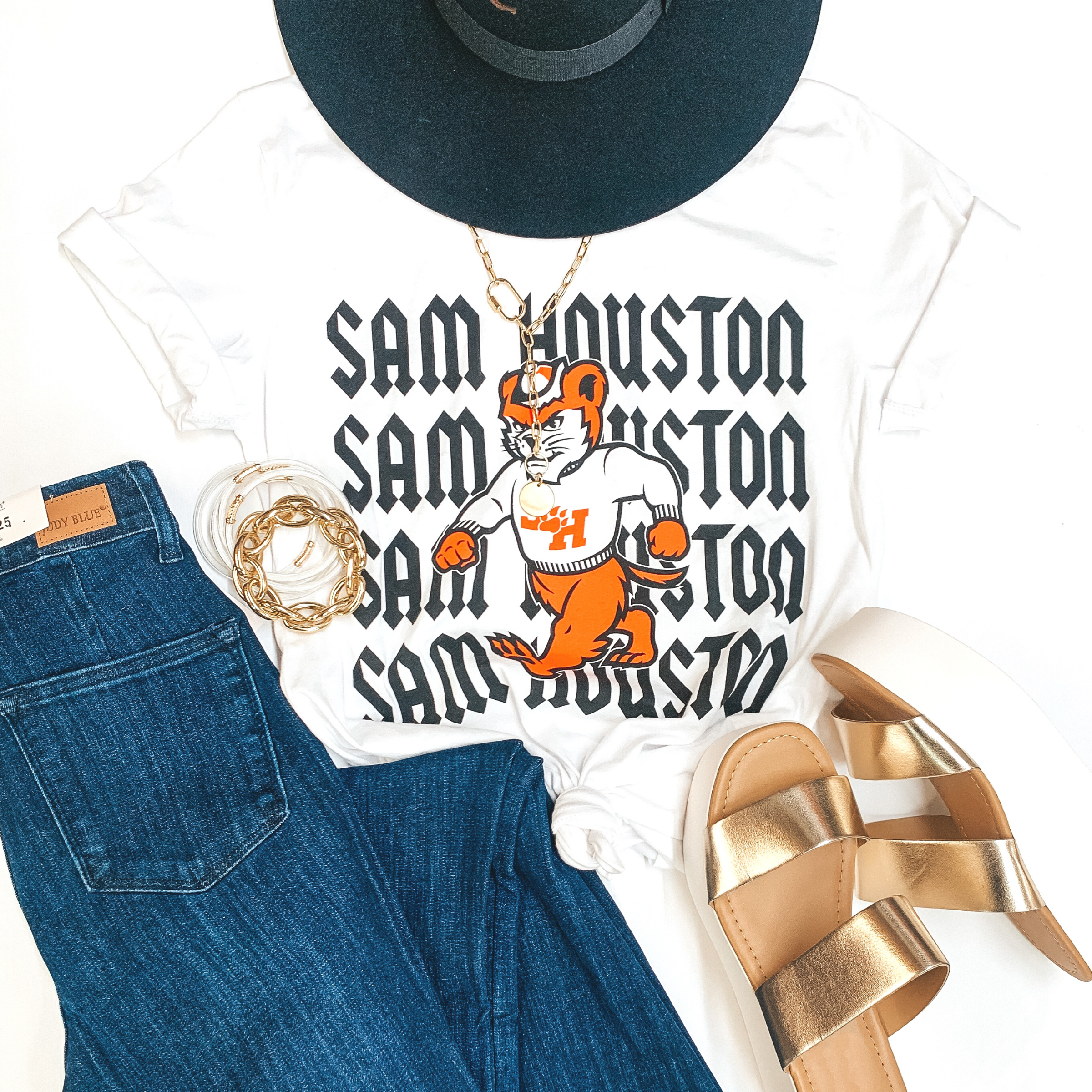 A white graphic tee with a bearkat that says "Sam Houston" in the background. Pictured on a white background with jeans, gold wedges, gold jewelry, and a black hat.