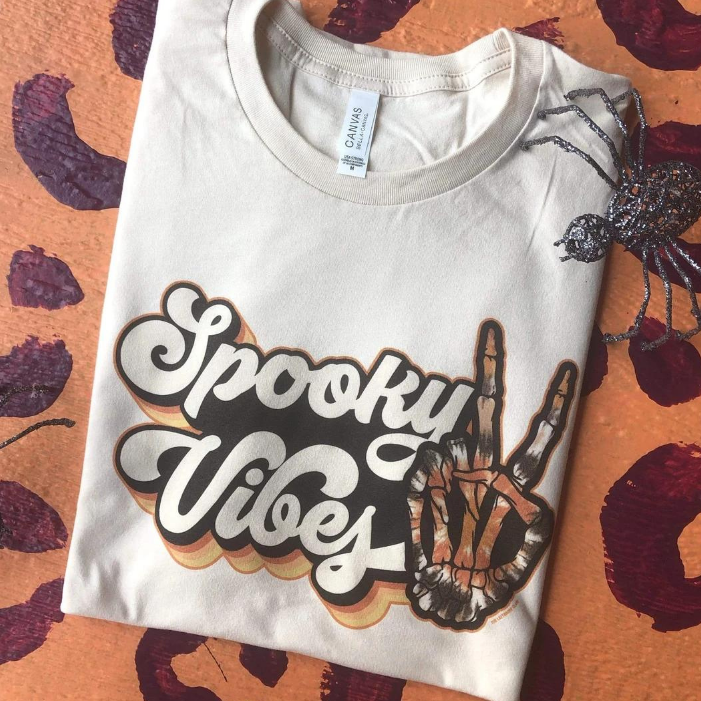 cream graphic tee that reads "spooky vibes" in a groovy font with a skeleton hand doing a peace sign hand gesture. Tee is laying on a leopard print backdrop with a fake spider as a prop. 