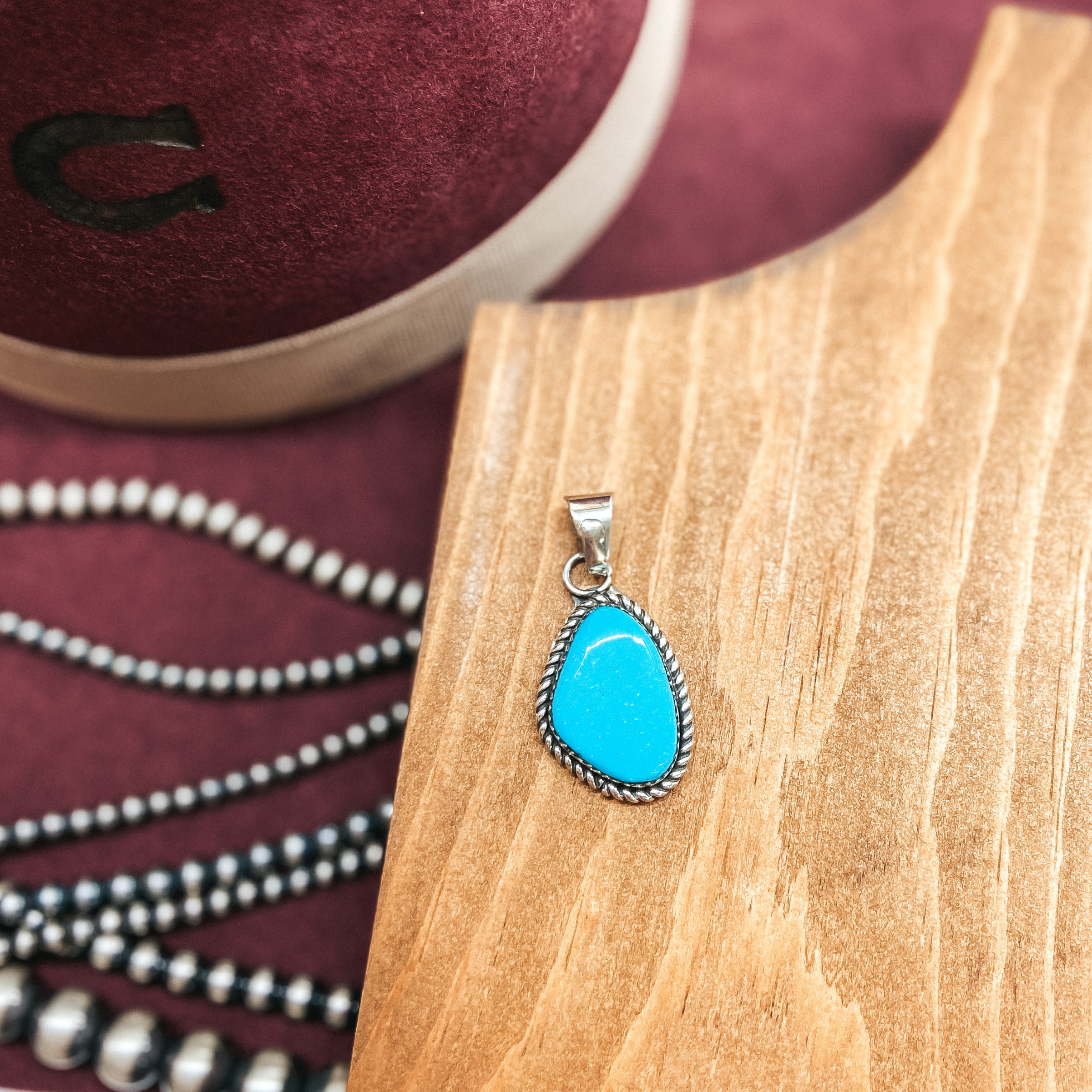 Turquoise stone pendant in silver rope setting. Pictured on wooden display with maroon hat and silver pearls.