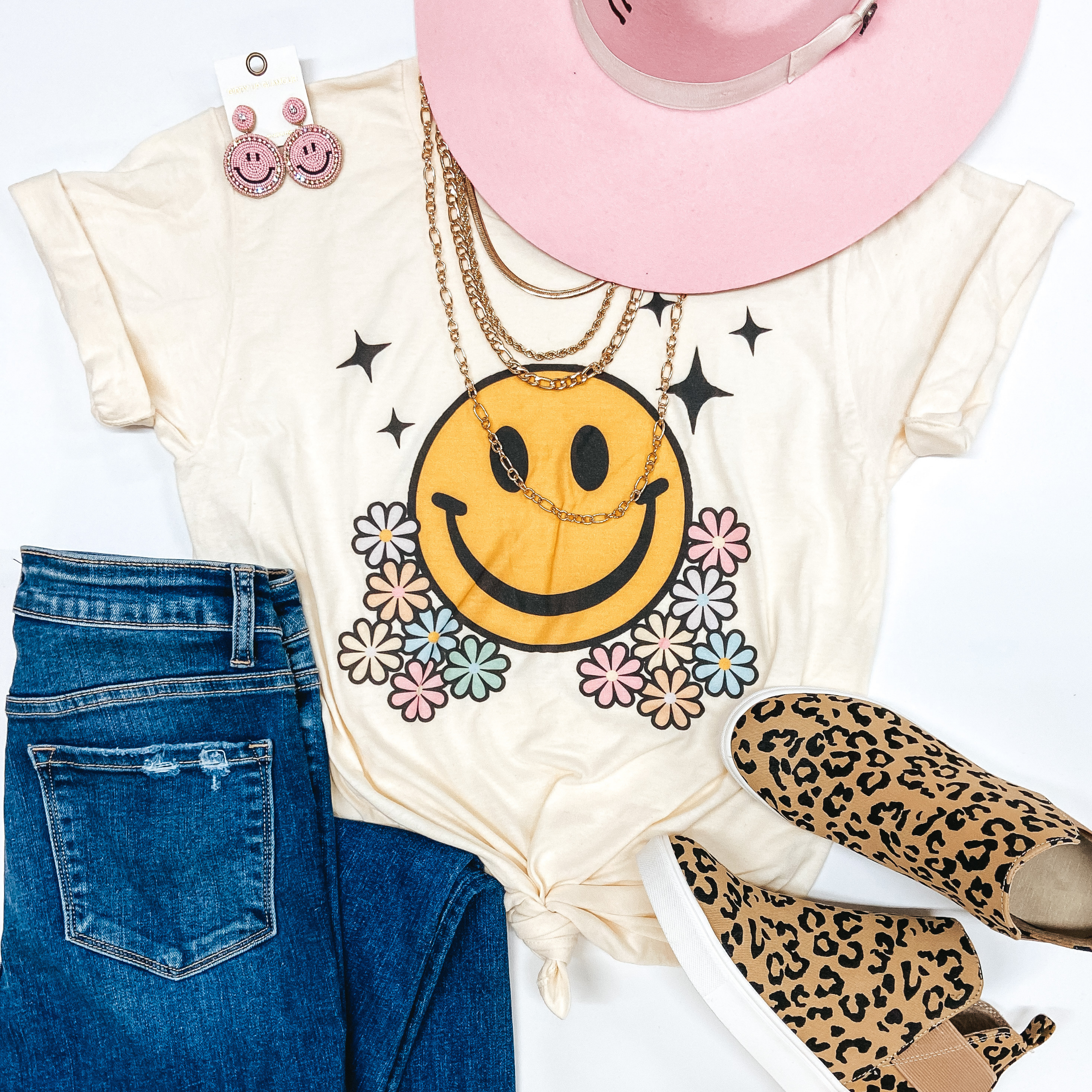 White graphic tee with a big yellow smiley face, sparkles, and bunches of pink, orange, and yellow flowers is styled on the group with a pink Charlie One Horse Hat, pink glittery smiley face earrings, medium wash jeans, and leopard print slide on sneakers. 
