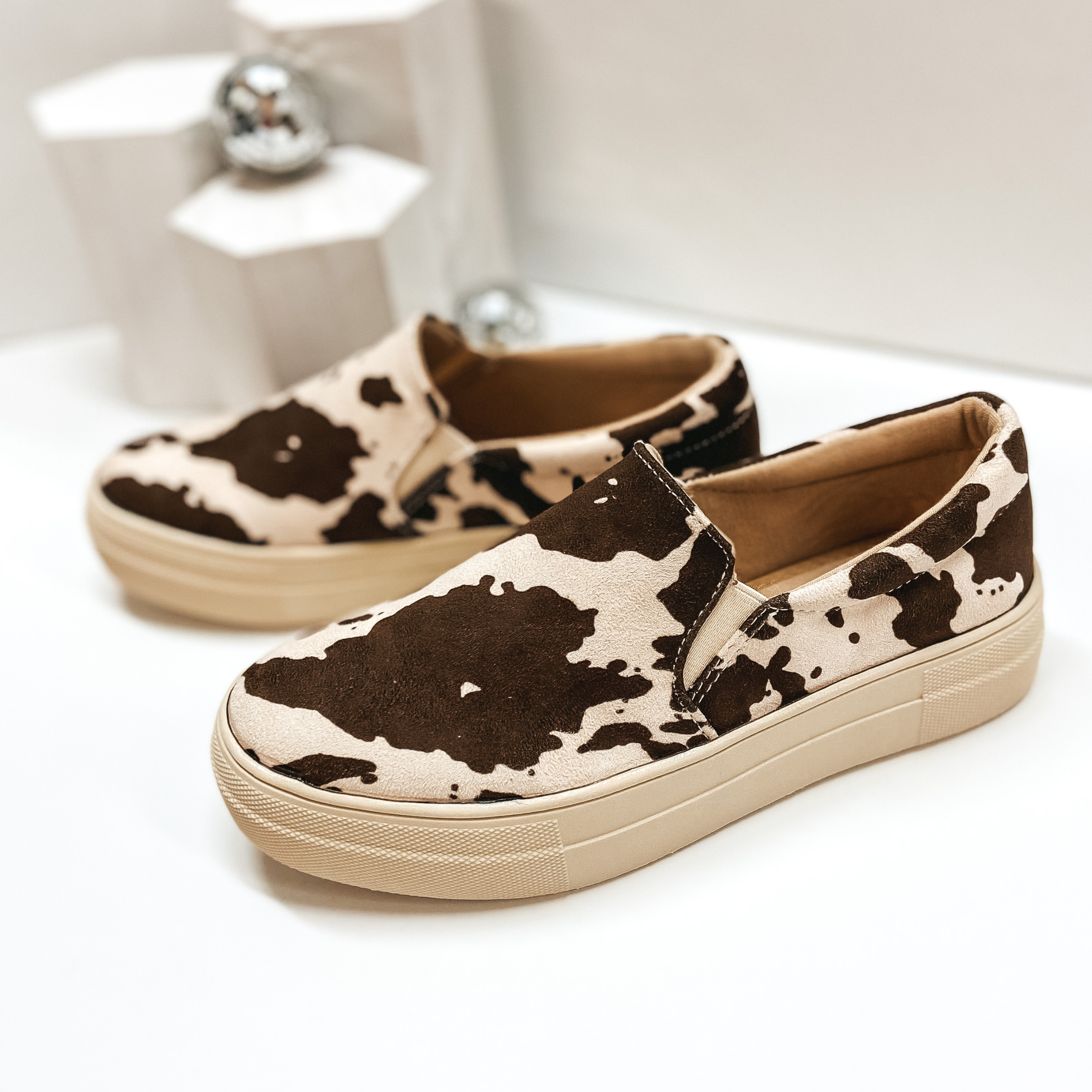 Slip on brown and ivory cow print sneakers with thick ivory sole. Pictured against a white background.