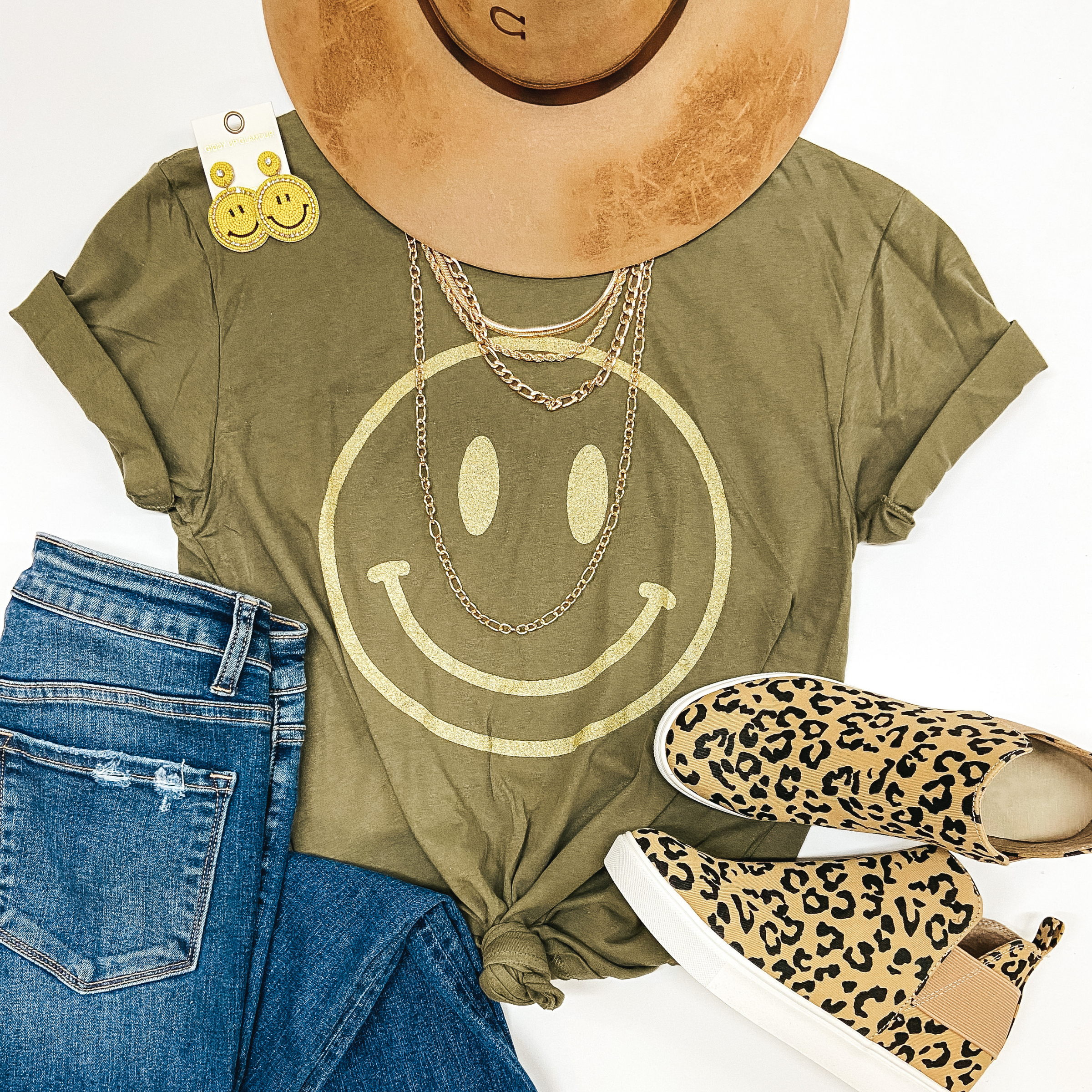 Olive green tee with a light green smiley face printed on it lies on a white background with a tan Charlie 1 Horse Hat, some necklaces, yellow glittery smily face earrings, leopard print slide on sandals, and medium wash jeans. 