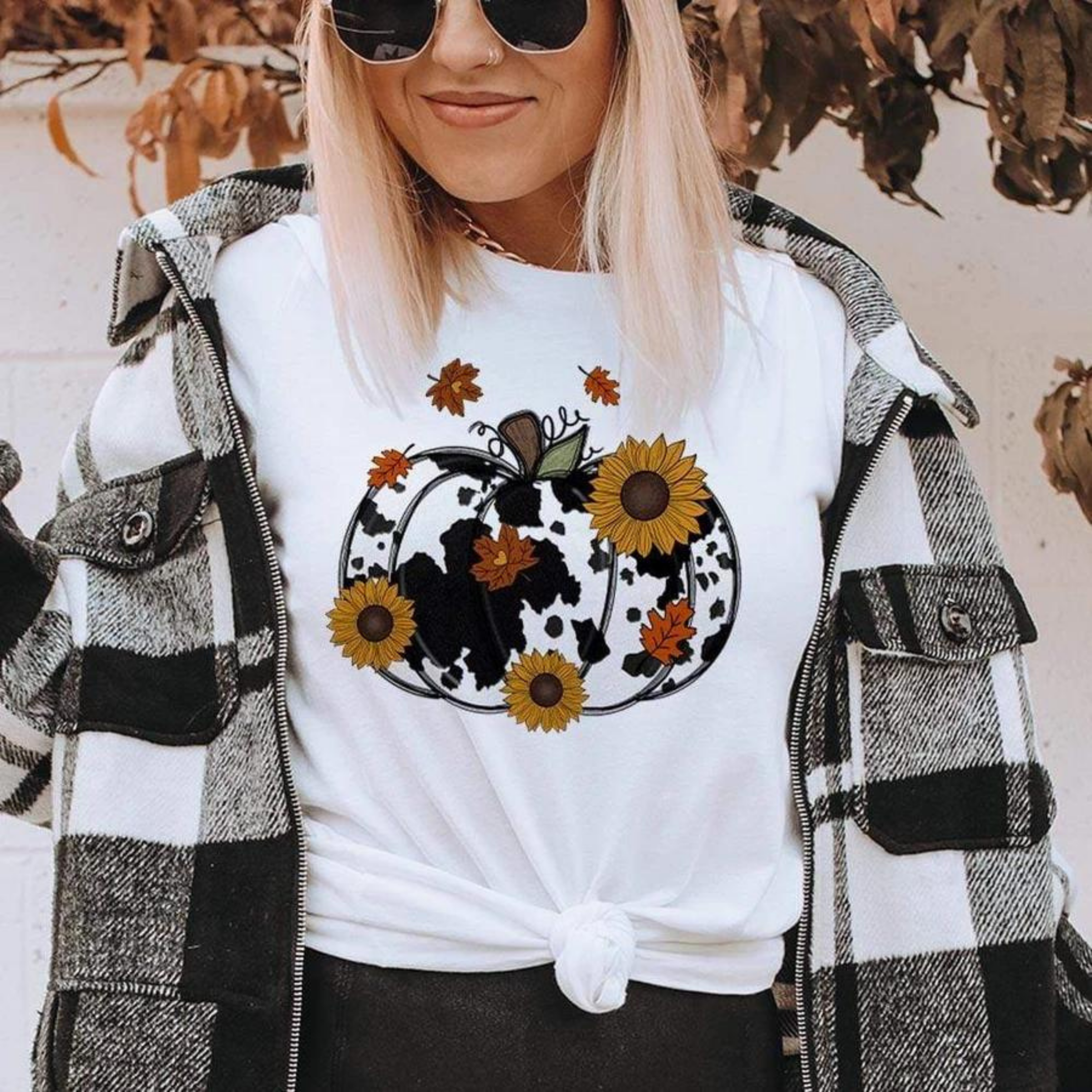 Model is wearing a white tee with a pumpkin graphic on it. The pumpkin is a cow print with sunflowers and leaves falling over top of the pumpkin. The model is also wearing a black and white buffalo plaid shacket and black pleather leggings. 