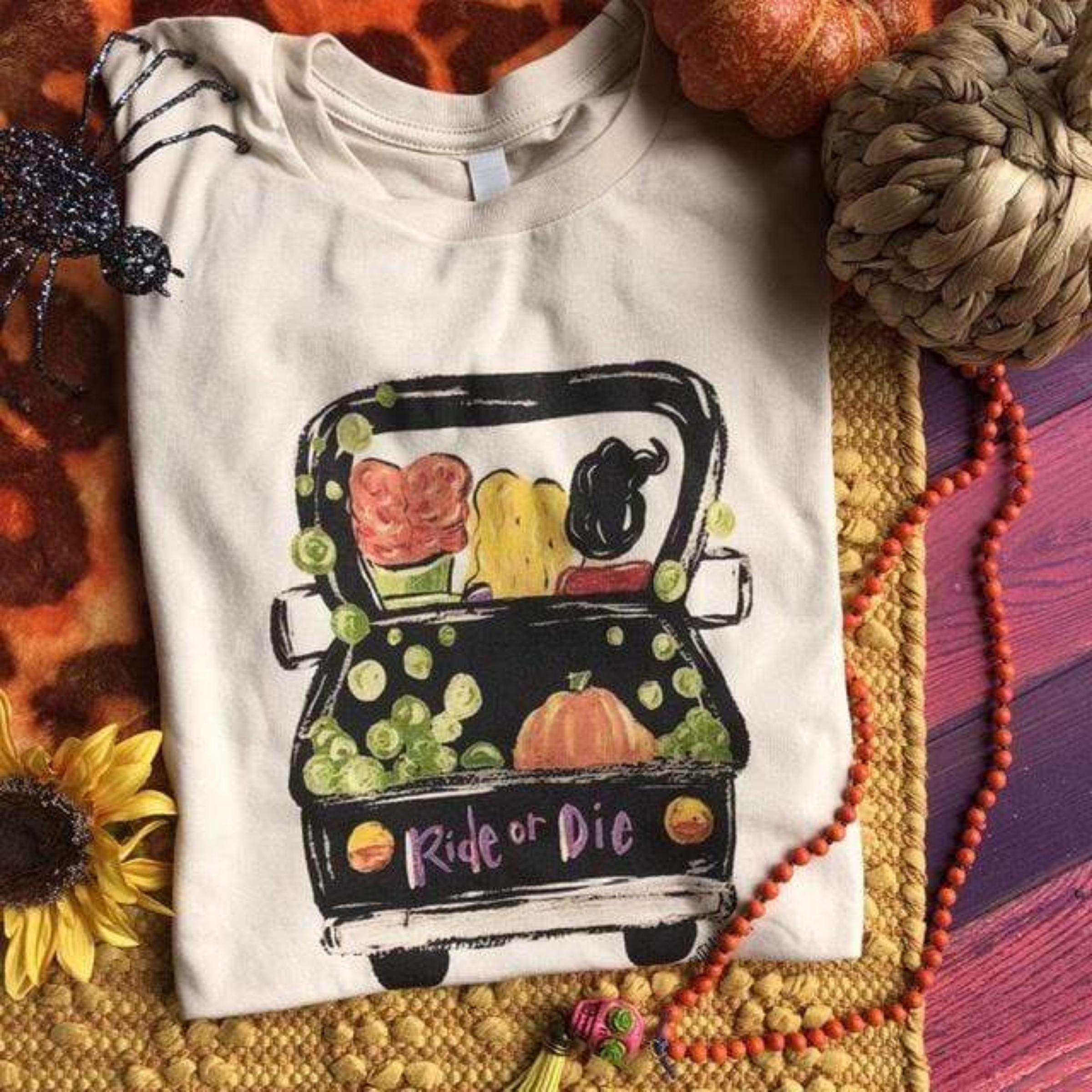 This is a cream t-shirt with a black truck on it with 3 girls inside the truck. On the back of the truck its says ride or die with a pumpkin and green gourds in the bed of truck. There is two decorative pumpkins, a glittered spider and a sunflower in the background of this picture.