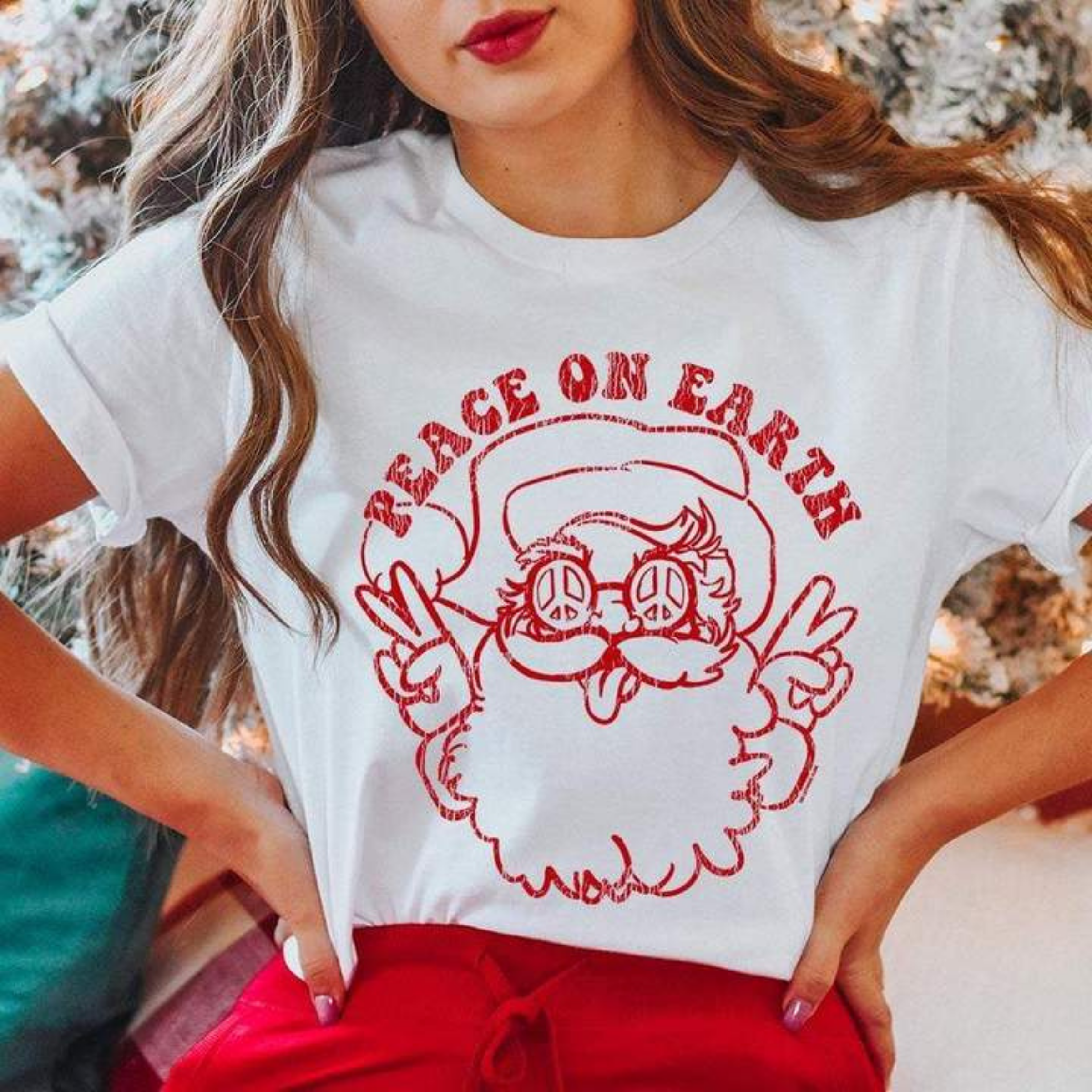 This white tee includes a crew neckline, short sleeves, and a graphic that says "Peace on Earth" and a hippy Santa Clause under the saying all in red. This is shown modeled with rolled sleeves and a pair of red pants. 