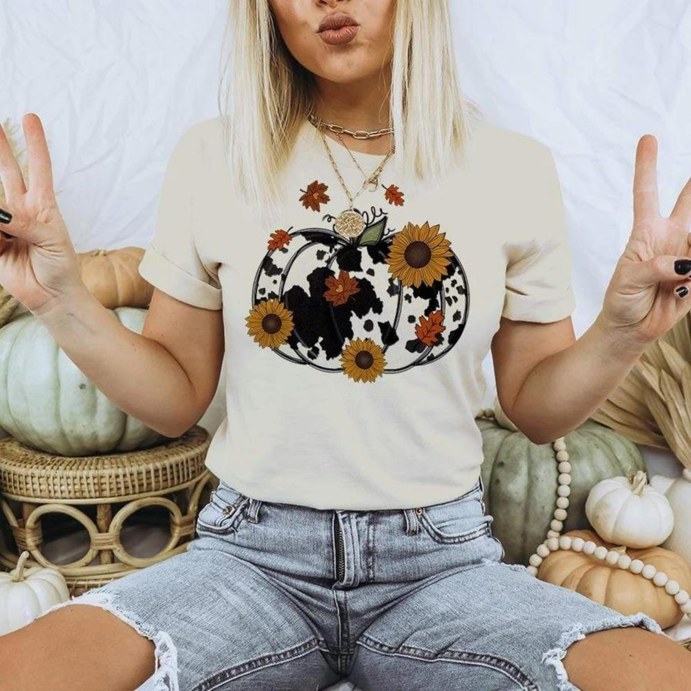 Model is wearing an ivory tee with a pumpkin printed on it. The pumpkin is filled with a cow print and has sunflowers and leaves floating over top of the design. Model is also wearing high waisted ripped jeans. 