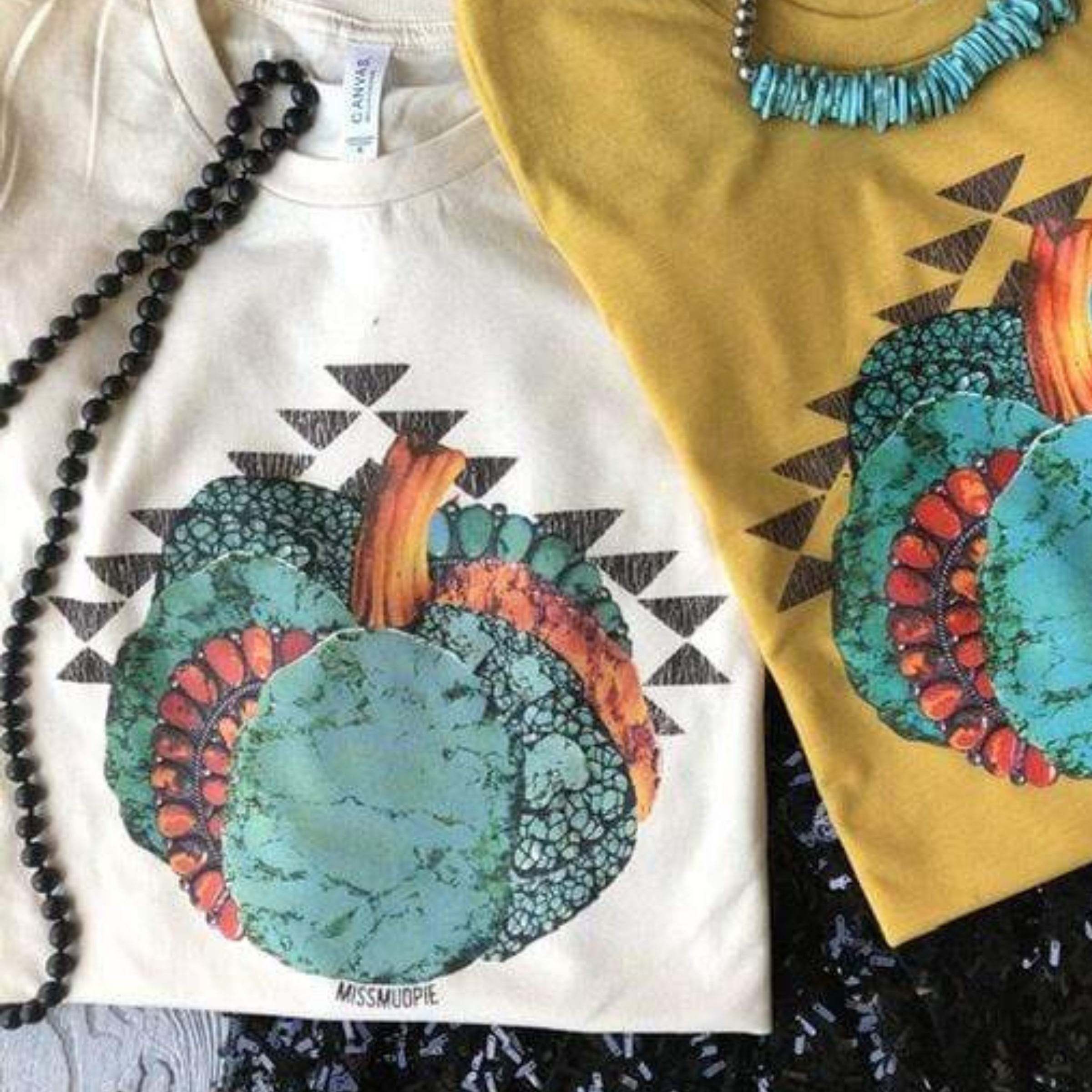 This is a cream graphic tee with a turquoise pumpkin. There is a red accent piece of jewelry in the side of the pumpkin. There are multiple many triangles surrounding the pumpkin. This top is paired with a long black beaded necklace.