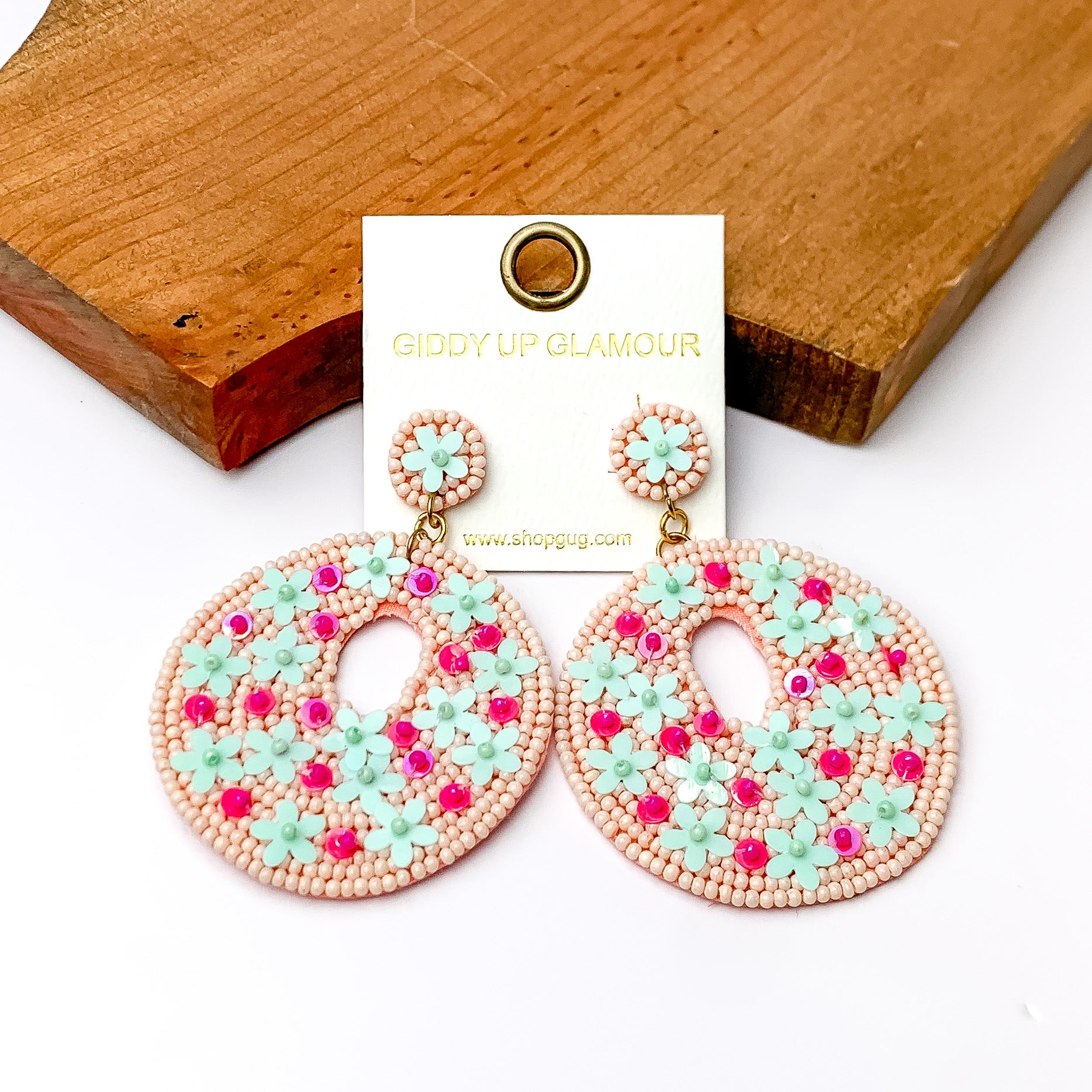 Light pink beaded circular drop earrings with pink and blue designs. Pictured on a white background with a wood piece at the top.