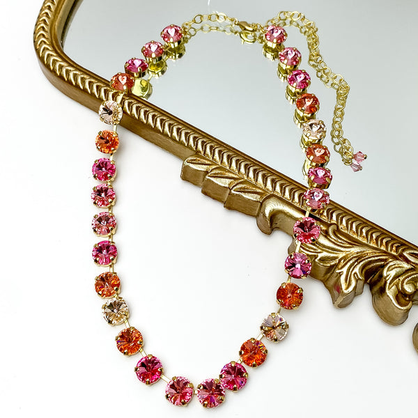 Mix of pink round crystal necklace. This necklace has a gold setting. This necklace is pictured partially laying on a gold mirror on a white background.    