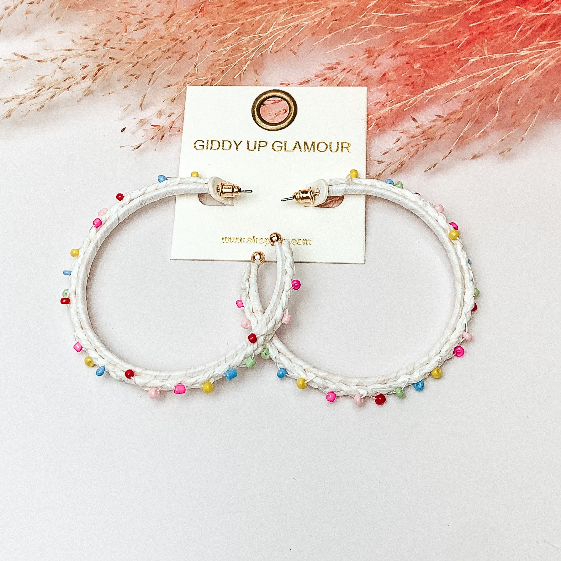 Pictured are raffia braided hoop earrings in white with colorful beads. They are pictured with a pink feather on a white background.