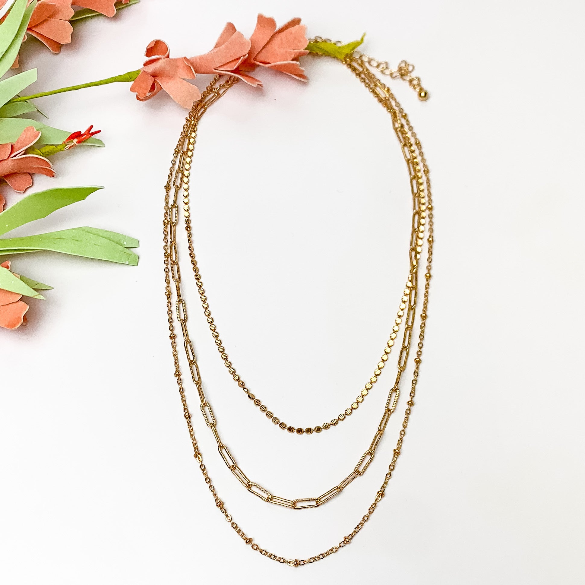Gold tone three layered chain necklace. Pictured on a white background with flowers to the left.