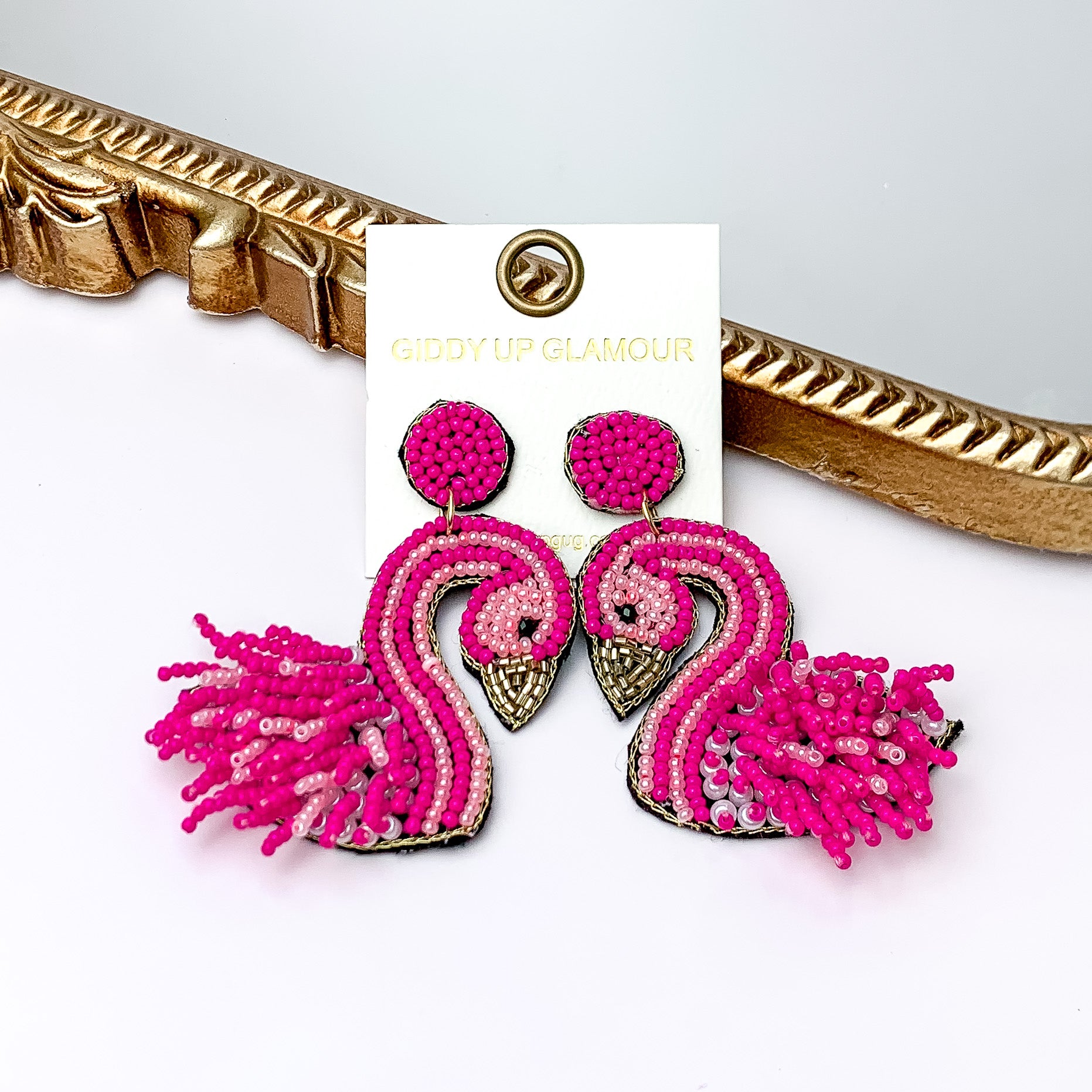 These earrings include circle studs with fuchsia beads and a gold outline. On the flamingo shaped beaded pendant there is beaded fringe detailing. These earrings are pictured leaning against a gold mirror on a white background.