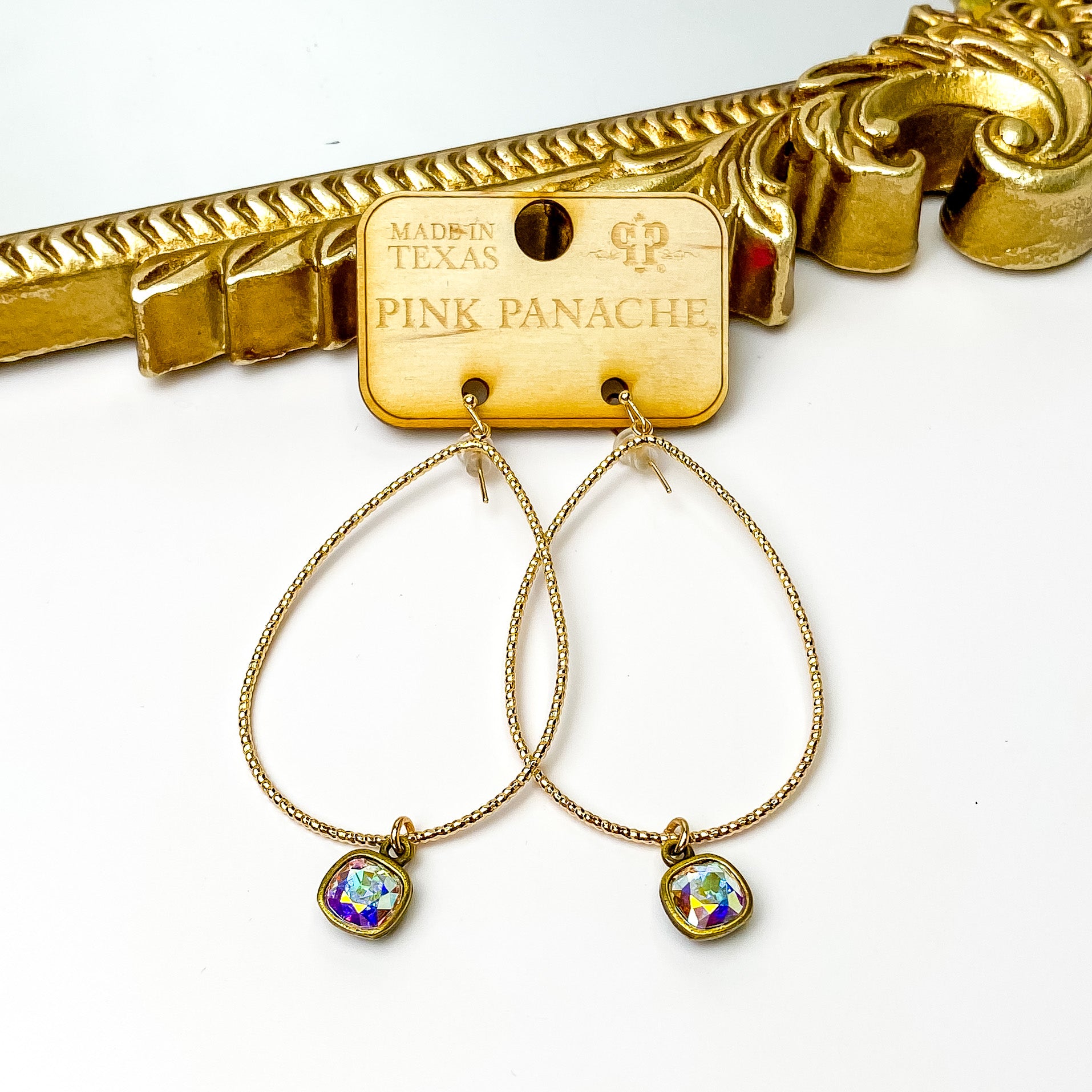Gold teardrop earrings with hanging ab cushion cut crystal charm. These earrings are pictured on a wood earrings holder in front of a gold mirror on a white background. 
