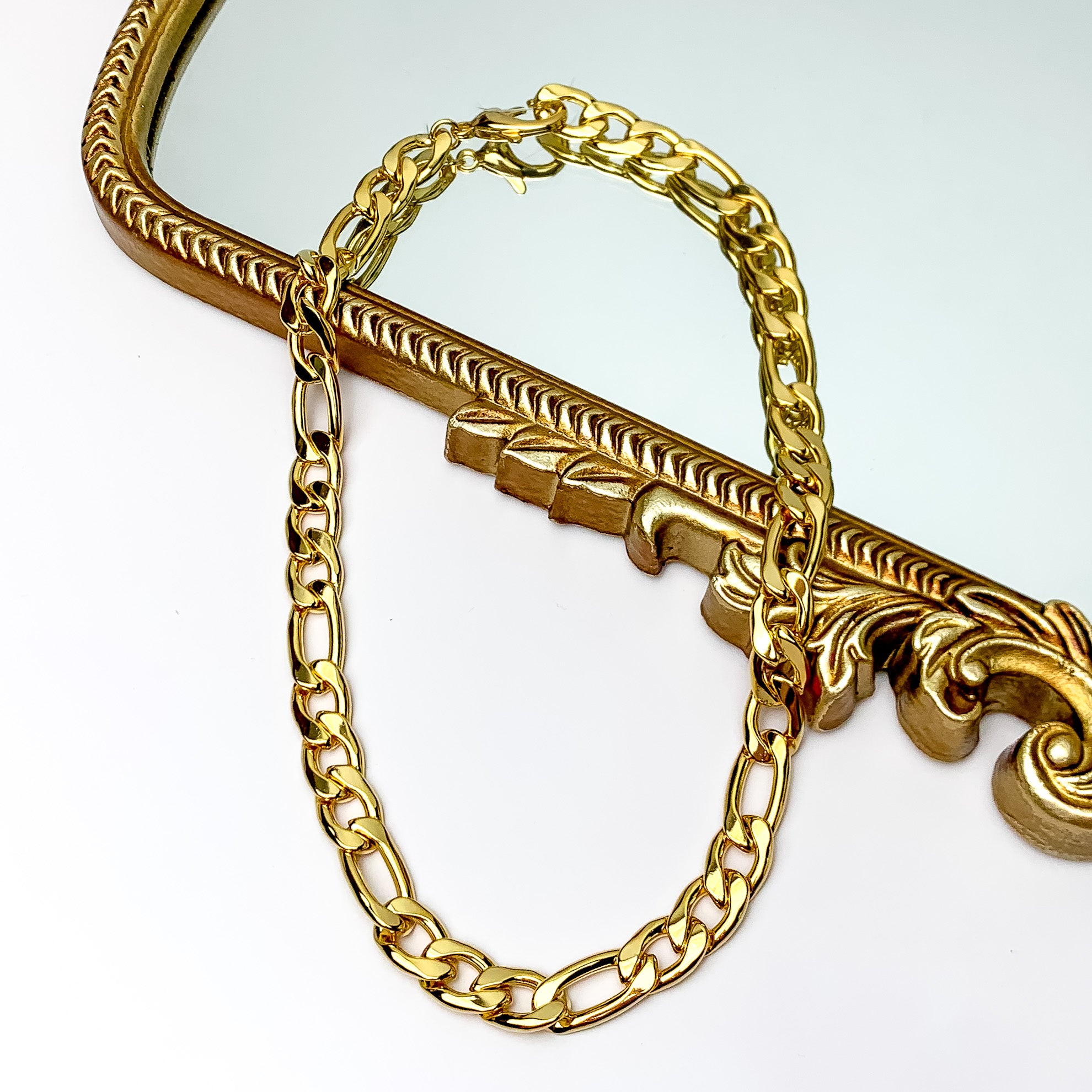 Pictured is a gold, curb chain necklace. This necklace is pictured partially laying on a gold mirror on a white background.  