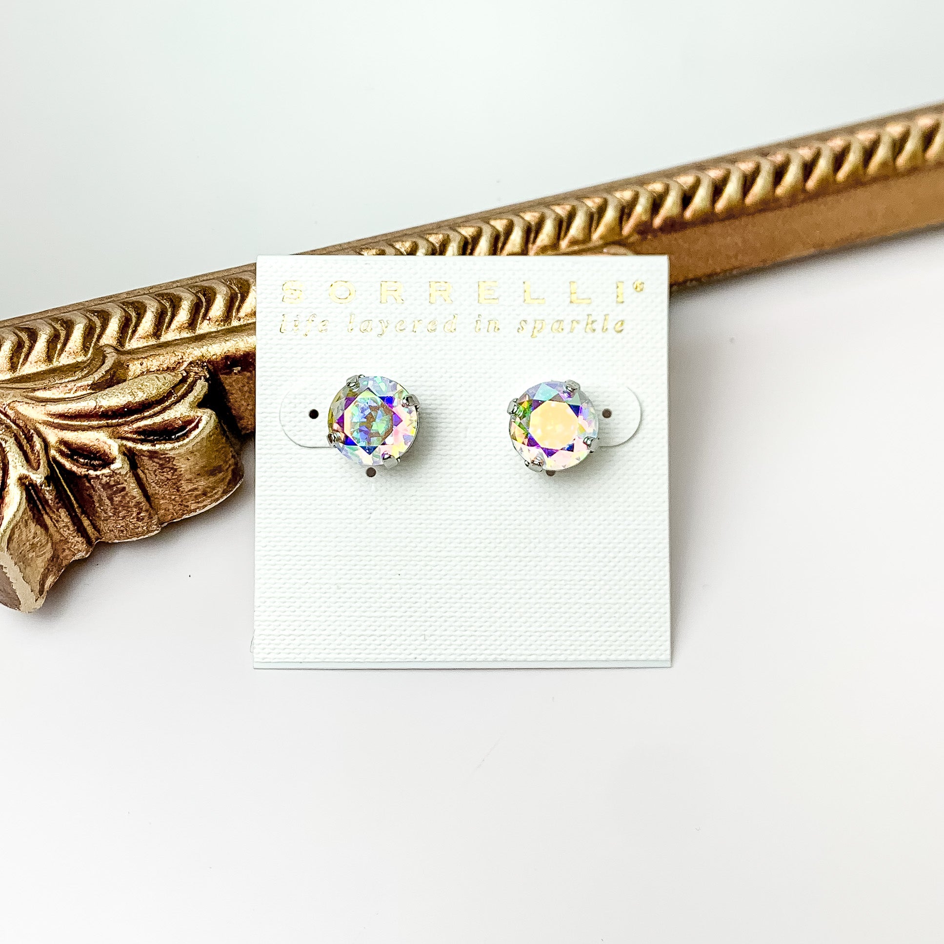 Pictured are round, ab crystal stud earrings with a silver backing. These earrings are pictured in front of a gold mirror on a white background.