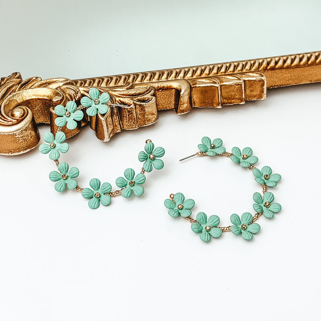 Gold hoop earrings with sage green flower charms outlining the earrings. These flowers have a gold bead center. One hoop is pictured laying against a gold mirror while the other hoop is laying flat on the white background. 