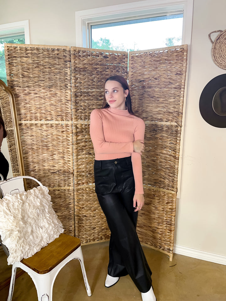 Model is wearing a high neck, ribbed long sleeve top in light pink. She is also wearing black leather pants with white boots, gold earrings and rings.