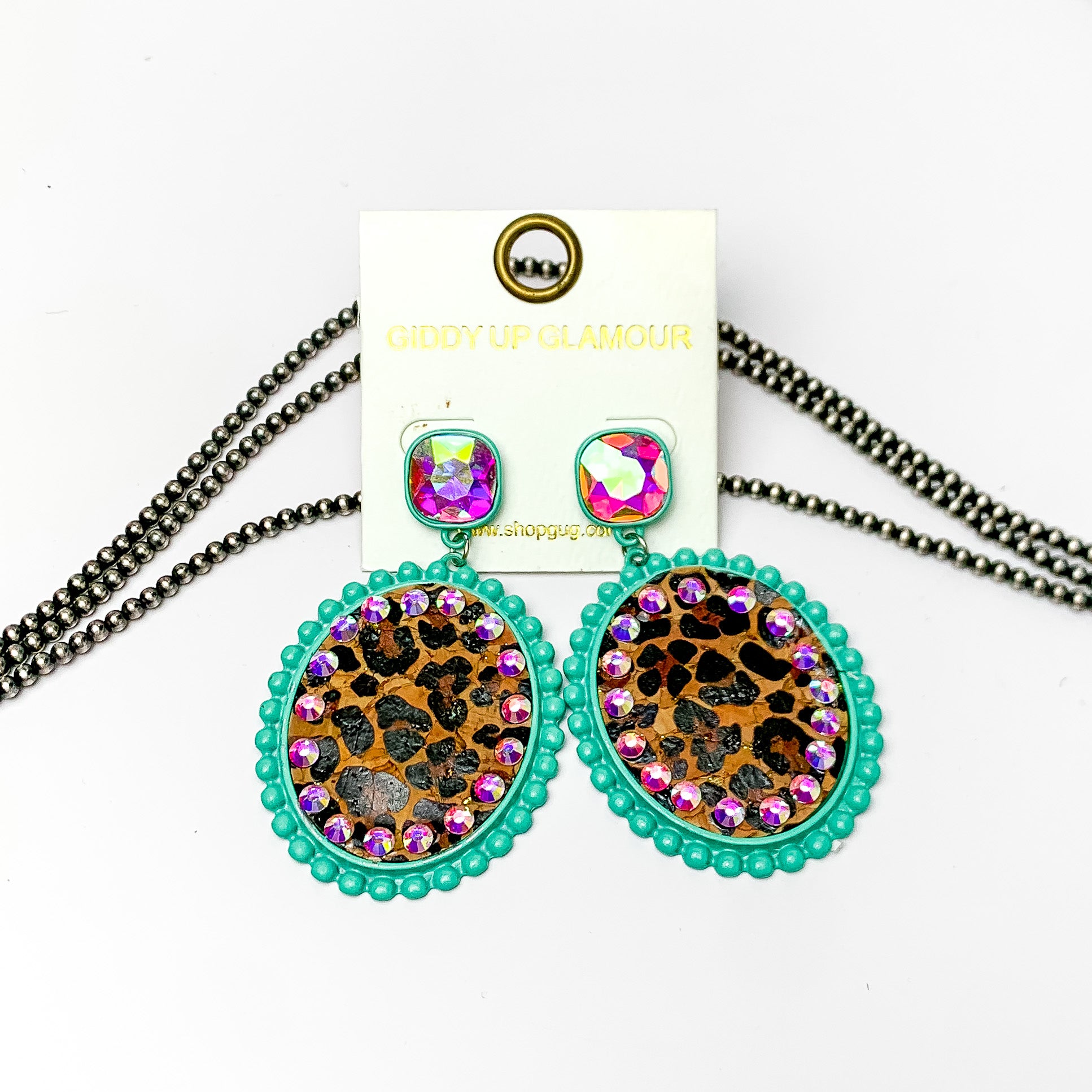 These earrings are oval shaped with leopard print inlay with the outline of the earing is turquoise but also has ab crystal going around the oval shape. These pair of earrings also have a ab crystal post style earring. Pictured on a white background with navajo pearls behind it.