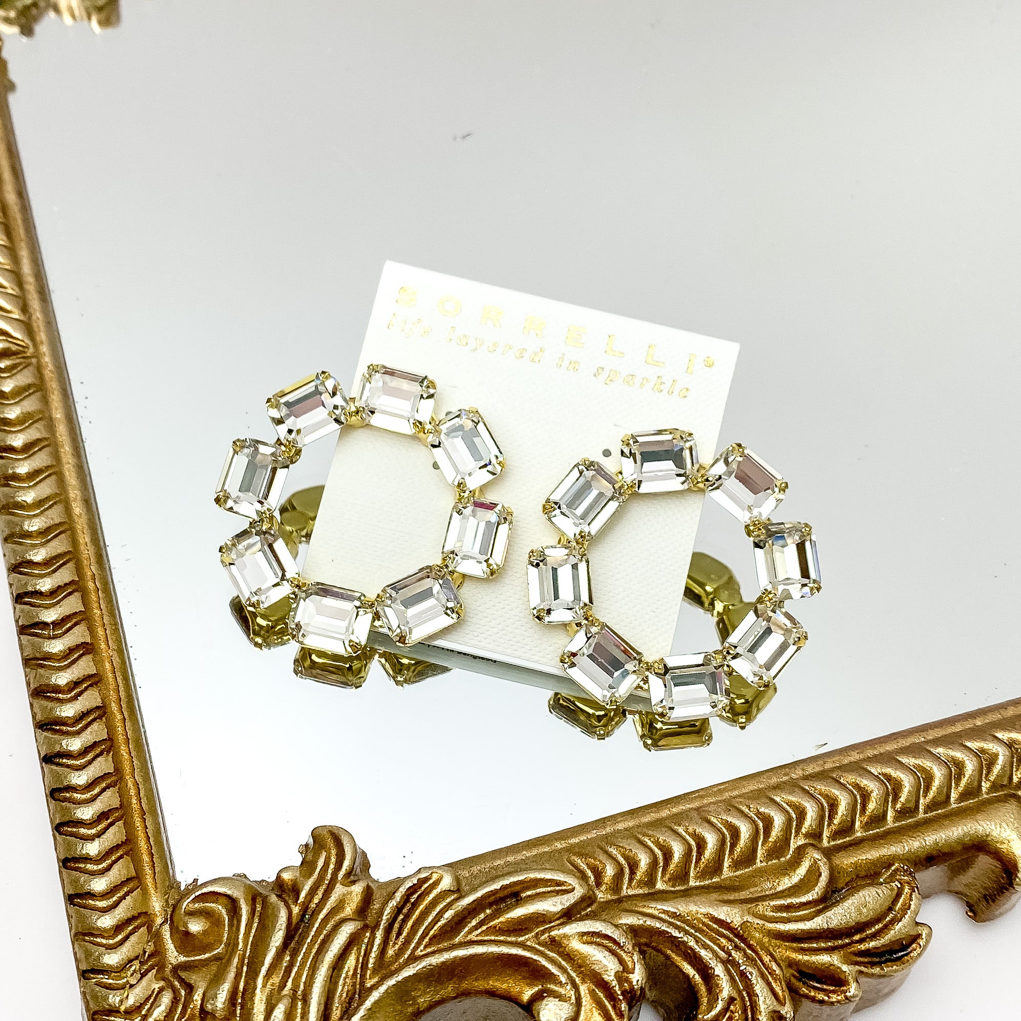 Eight rectangle clear crystals in the shape of an octagon post back earrings. These earrings have a gold setting and gold prongs to hold the crystals in place. These earrings are pictured on a gold mirror on a white background. 