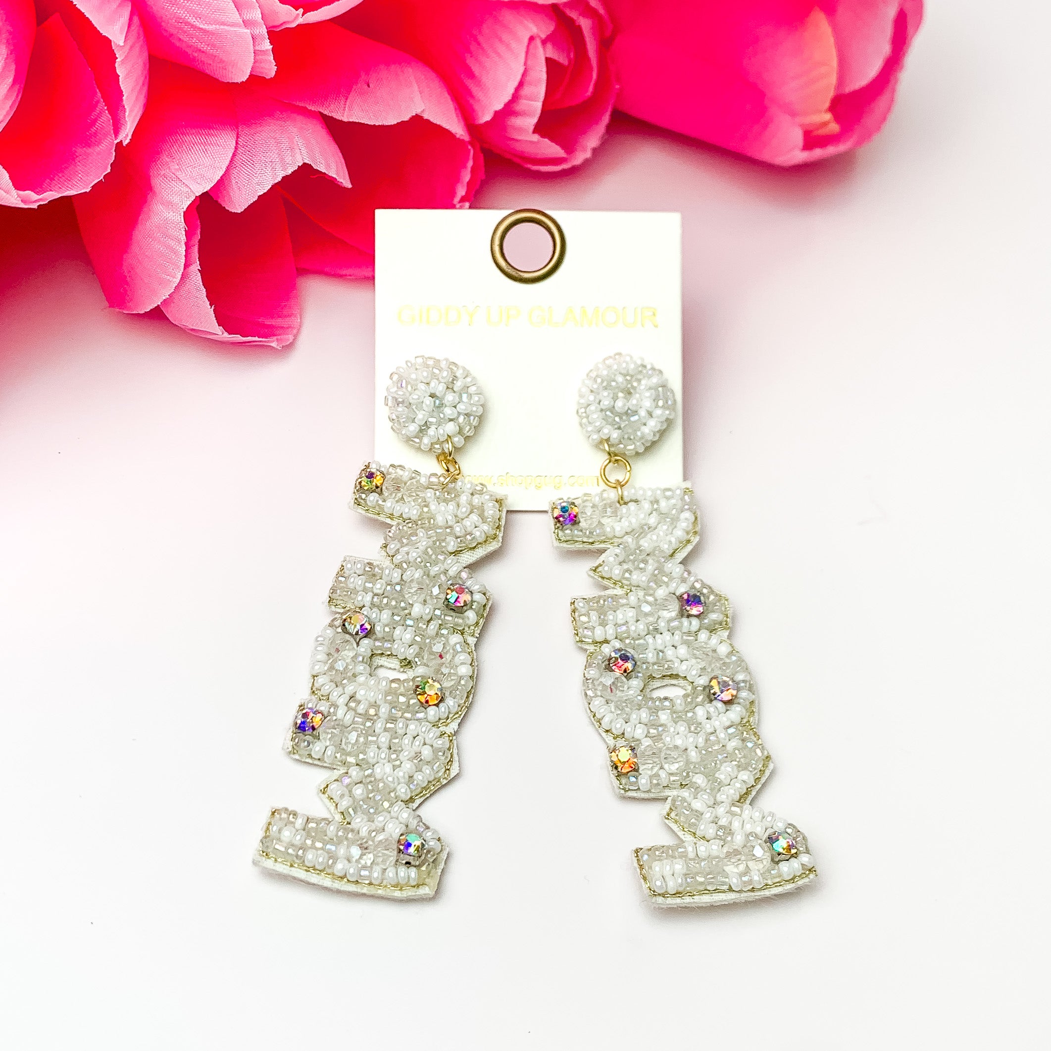 These earrings include white, circle, beaded post back earrings with a long dangle. This dangle spells out "MOM" in white beads with an inlay of ab crystals. These earrings are pictured on a white background with pink flowers.
