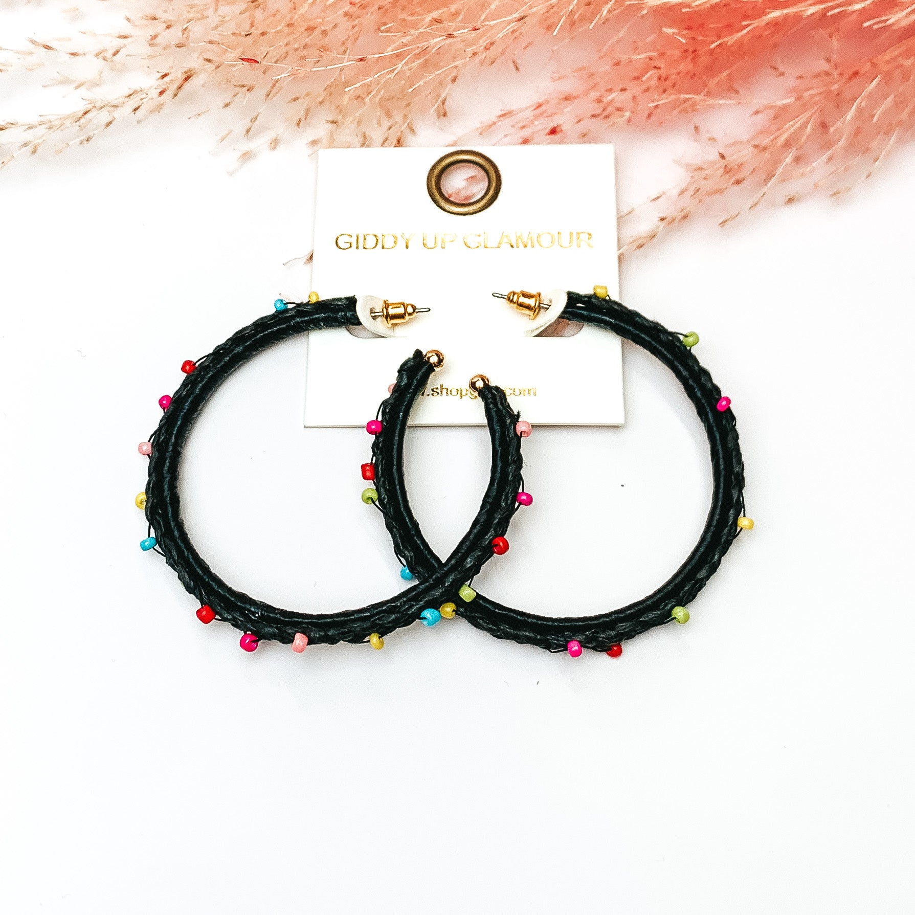 Pictured are raffia braided hoop earrings in black with colorful beads. They are pictured with a pink feather on a white background.