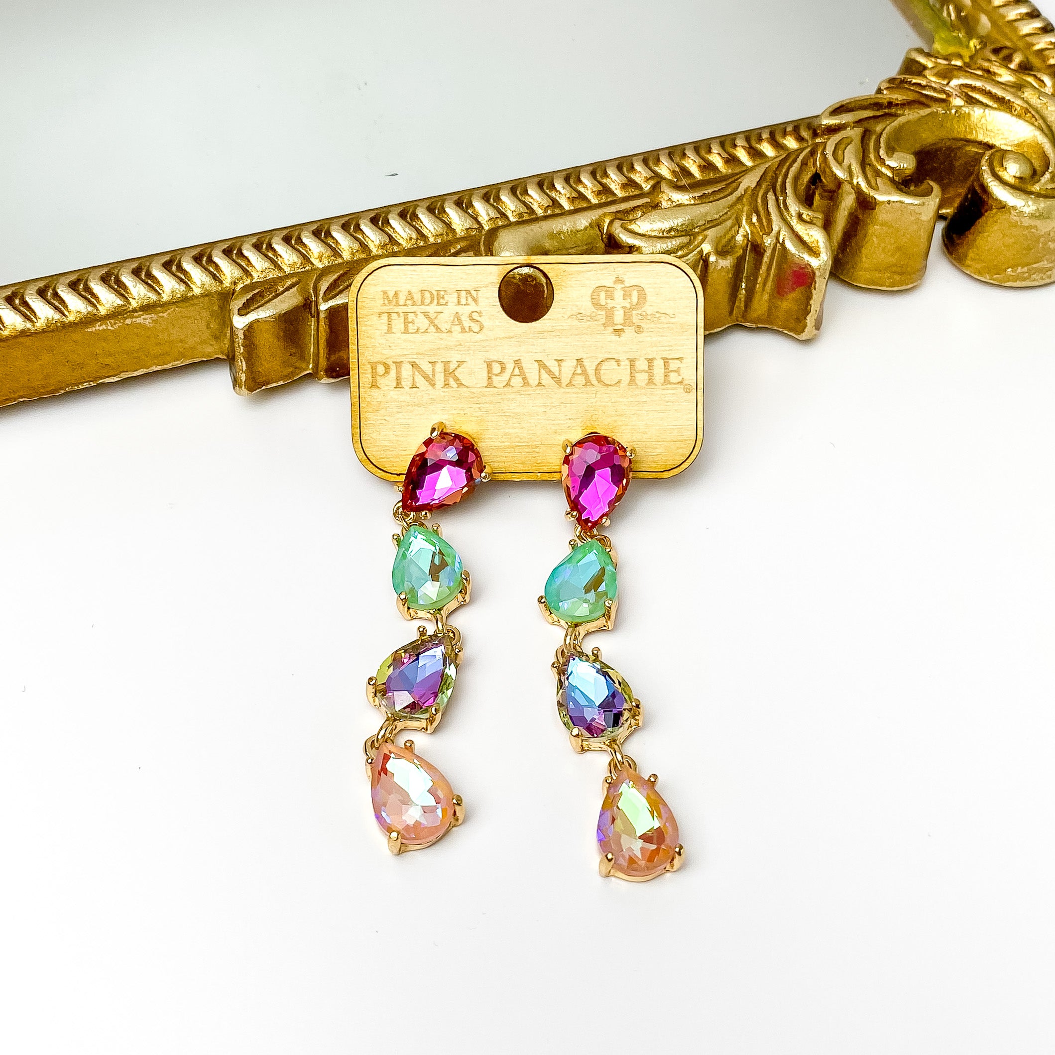 Four teardrop crystals that are linked together. The crystals include a fuchsia colored crystal, mint colored crystal, purple colored crystal, and light pink colored crystal. These earrings are pictured on a wood earrings holder in front of a gold mirror on a white background. 