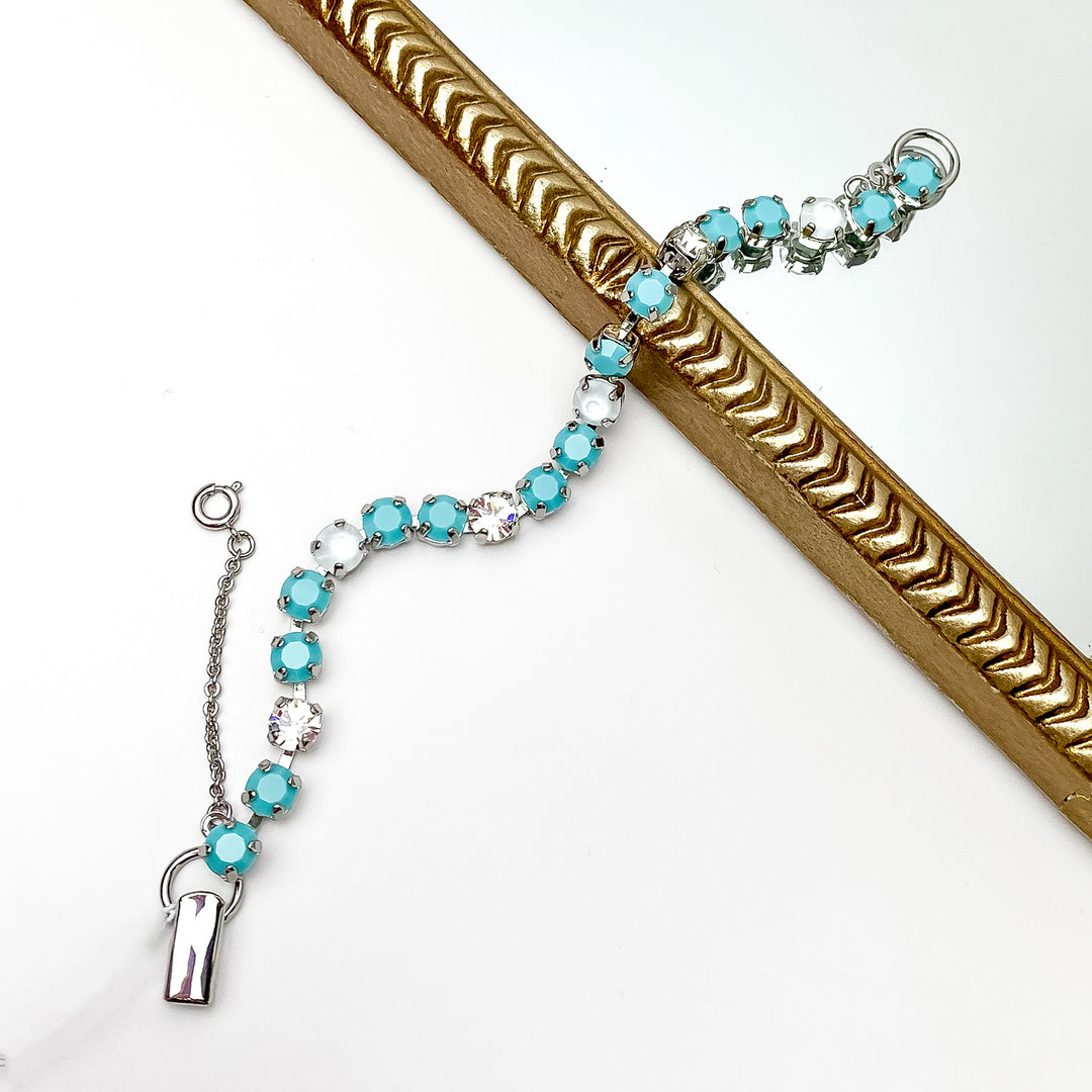 Silver tennis bracelet with light blue, white and clear crystals all around. Pictured on a white background and a gold frame through the middle.
