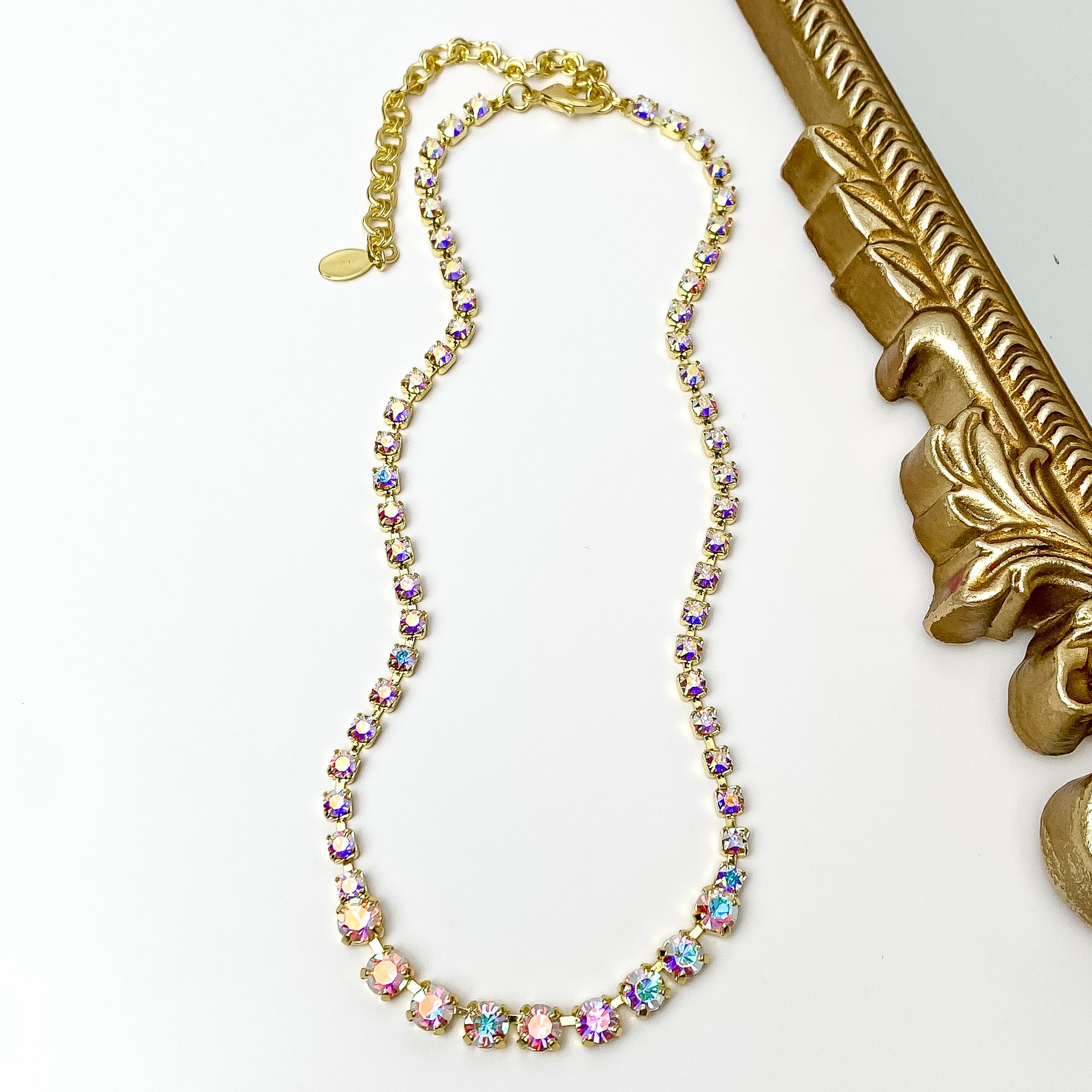 Pictured is a gold necklace with ab crystals. This necklace is pictured on a white background with a gold mirror on the right side.