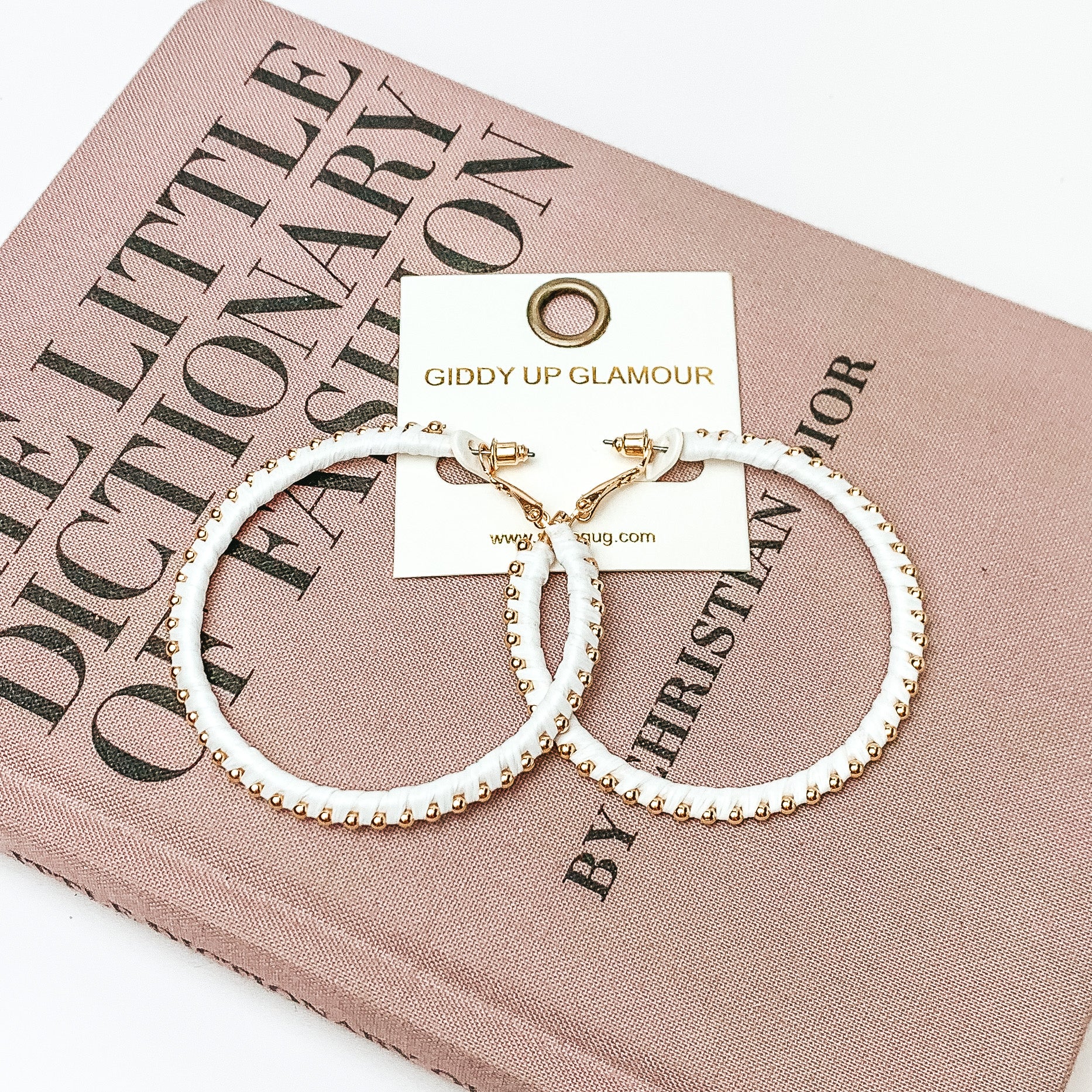 Pictured are circle pure white hoop earrings with gold beads around it. They are pictured with a pink fashion journal on a white background.