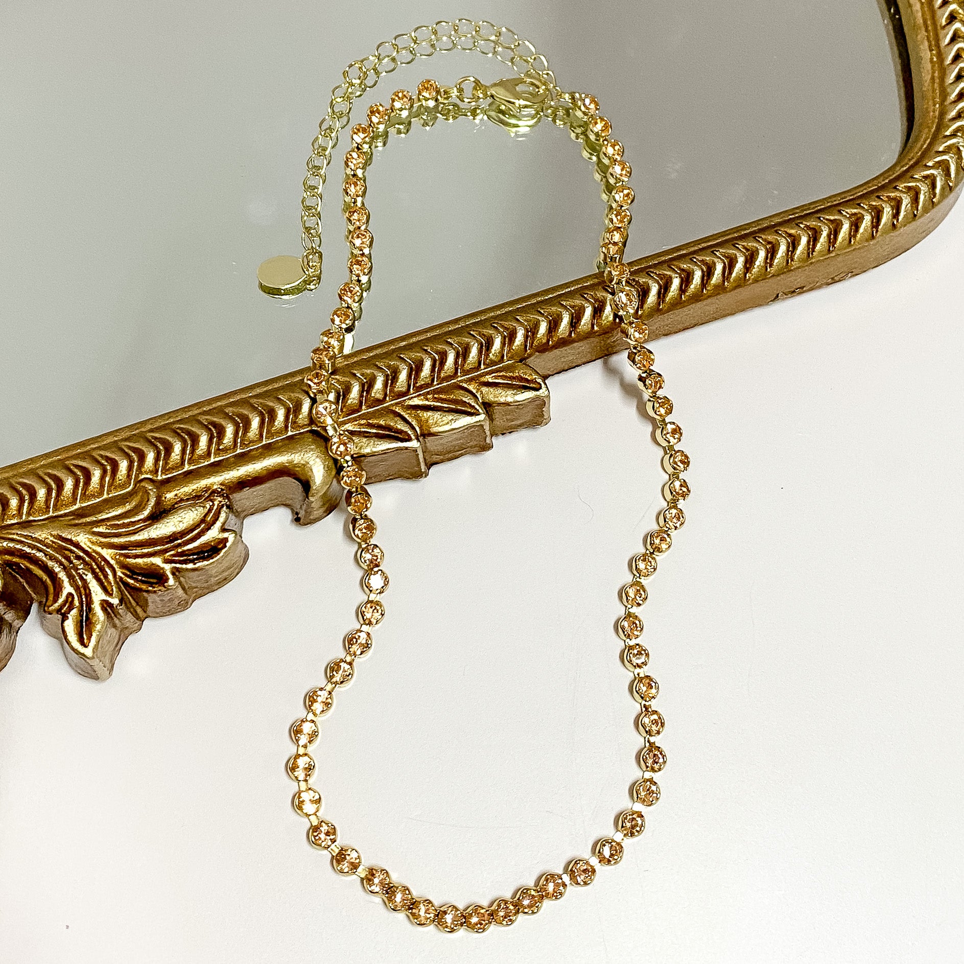 Pictured is a gold necklace with light beige crystals. This necklace is pictured partially on a gold mirror on a white background.